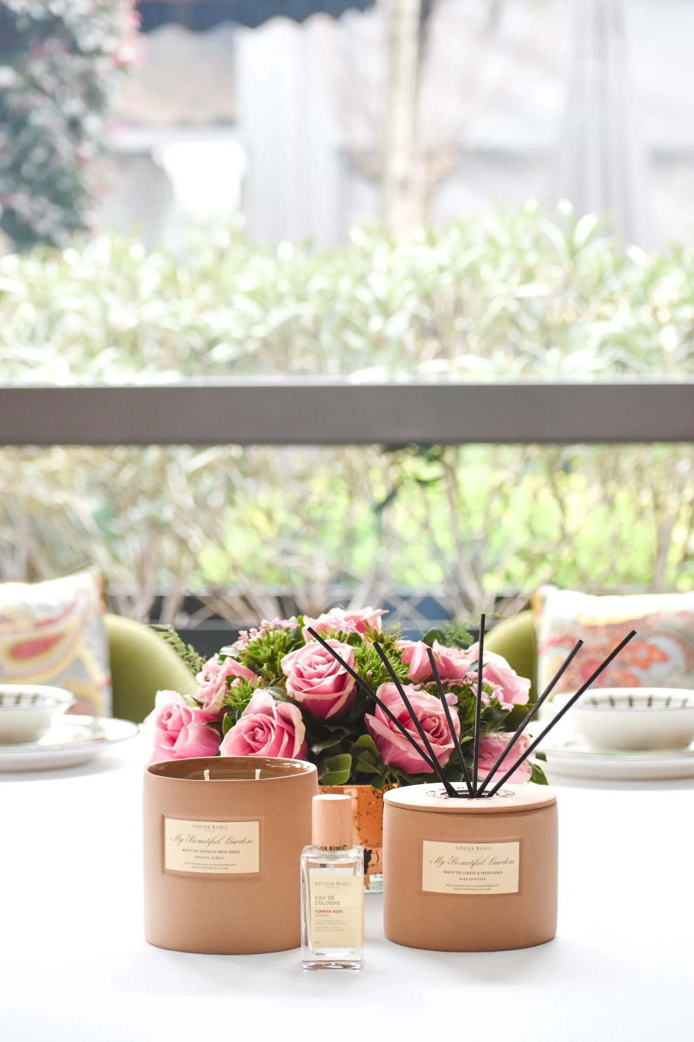 A table decorated with pink roses, candles, and perfumes, set near a window with a garden view.
