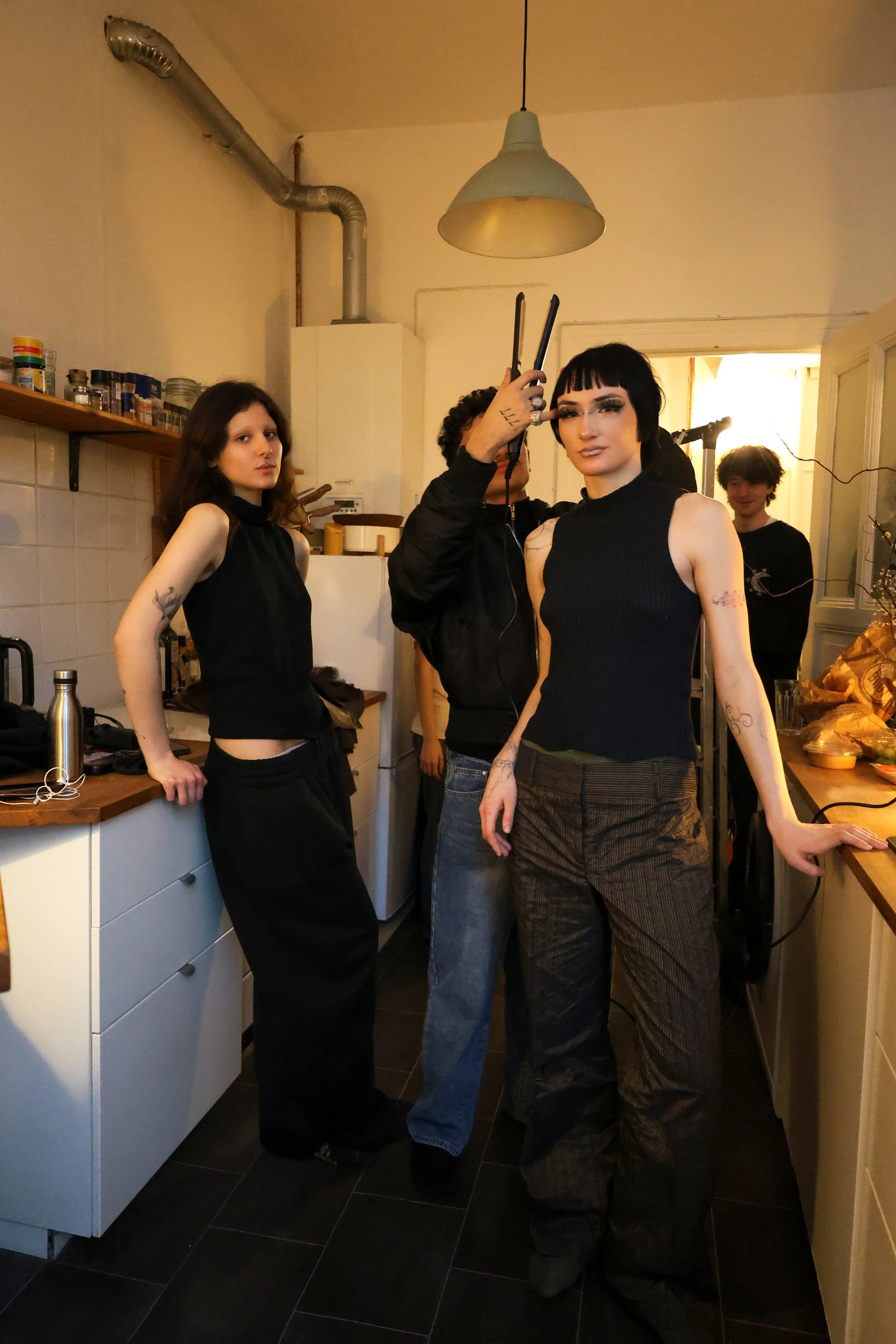 A group of four people standing in a kitchen with a modern industrial decor, including a hanging pendant light, cabinets, and a tiled backsplash. One person is holding a pair of scissors and looking at the camera, while the others are casually posed, some looking at the camera and others elsewhere.