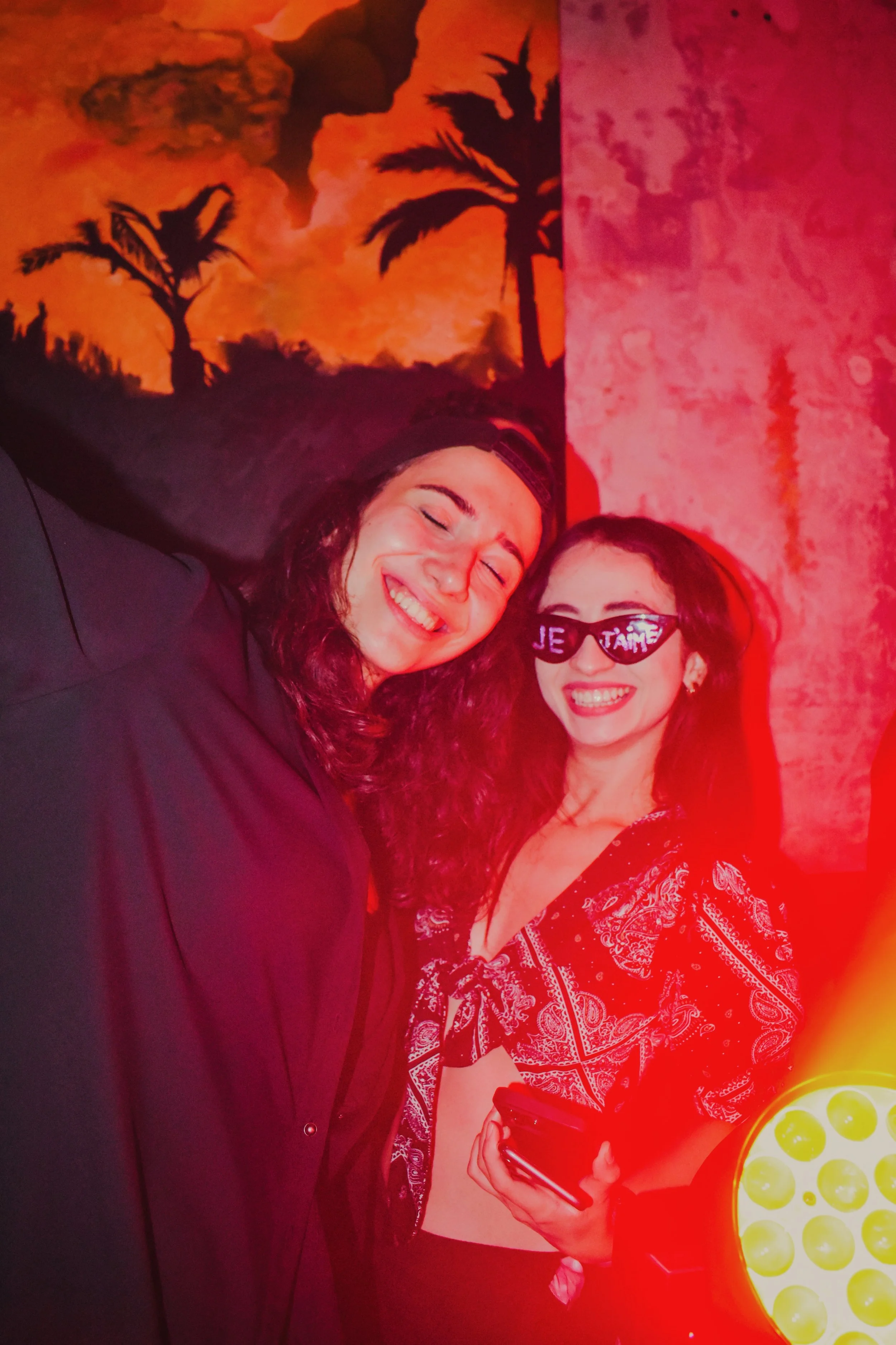 Two women smiling and enjoying themselves, one wearing sunglasses with 'JE T’AIME' written on them, in a colorful room with tropical palm tree artwork on the wall.
