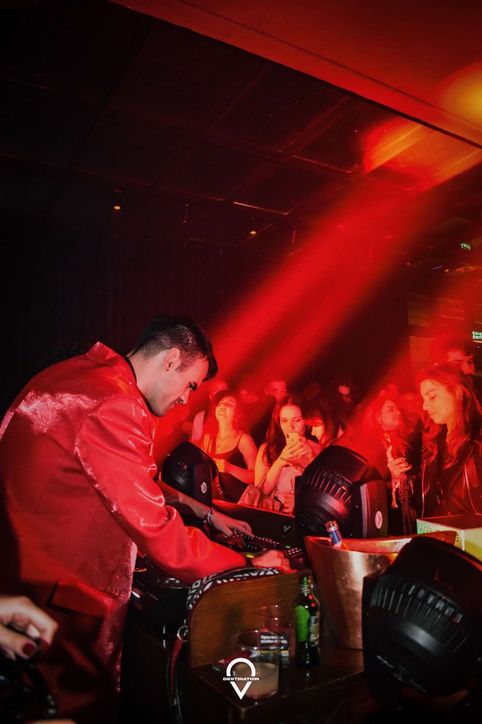 A DJ in a red jacket performs at a nightclub, surrounded by an audience with some taking photos. The scene is lit with red lights.