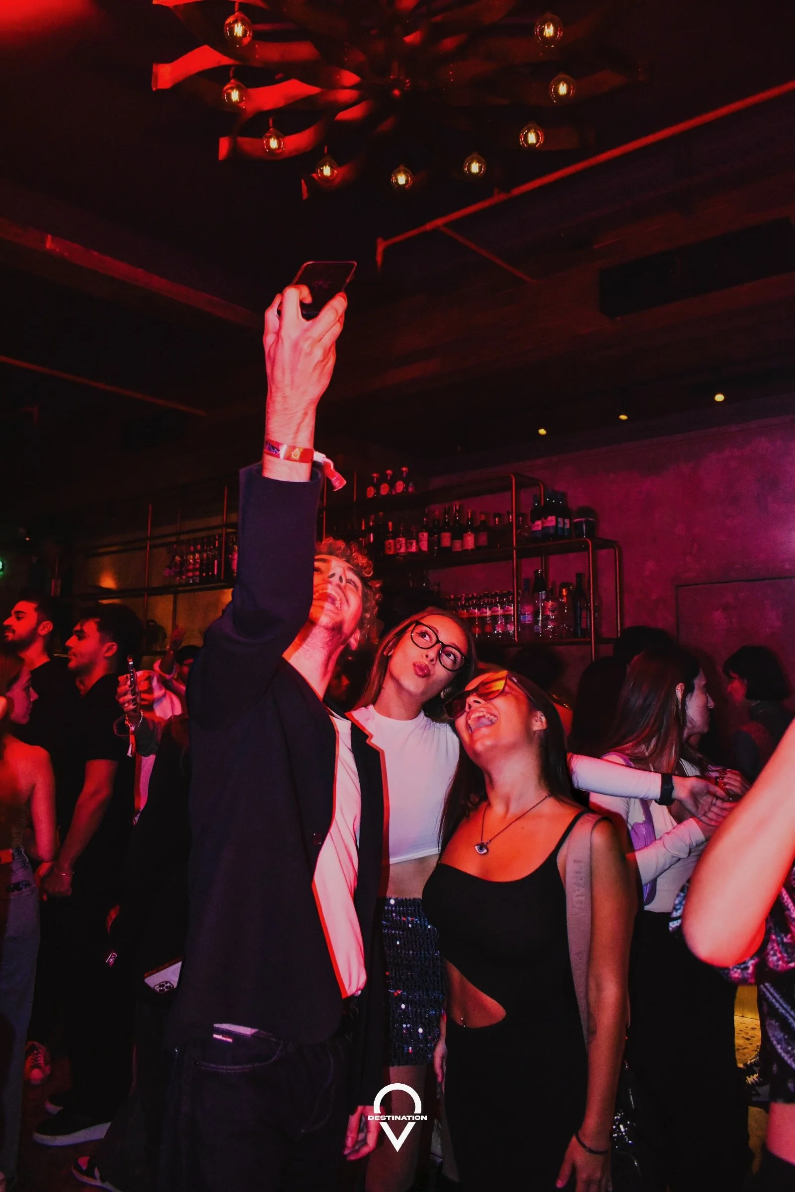 People dancing and posing for a selfie in a dimly lit nightclub with red lighting and shelves of bottles in the background.