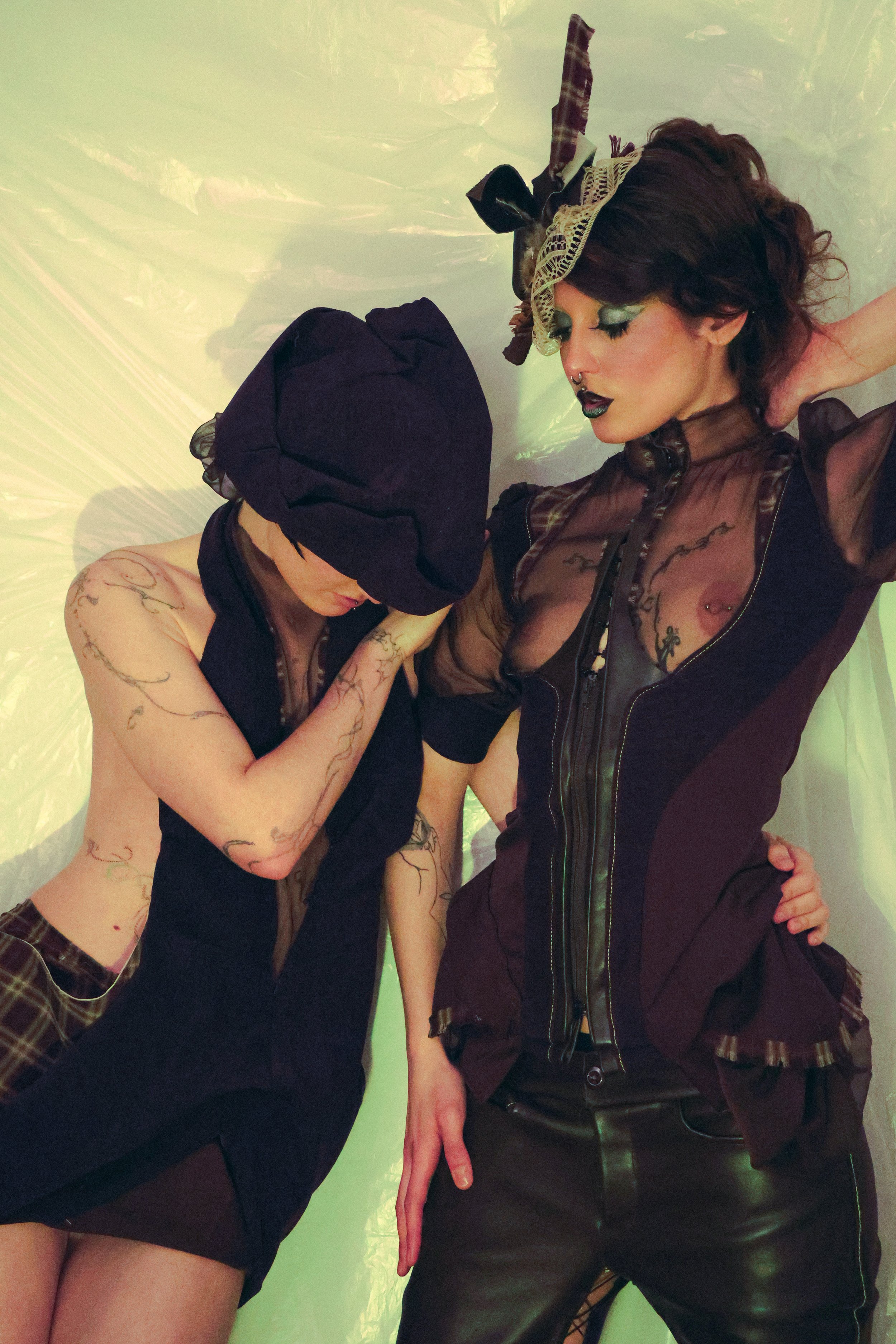 Two women in avant-garde fashion pose against a shiny, translucent backdrop. One wears a plaid skirt, a sheer top with tattoos visible, and a large dark hat covering her eyes. The other wears leather pants, a sheer top with tattoos visible, and a decorative headpiece with lace and fabric.