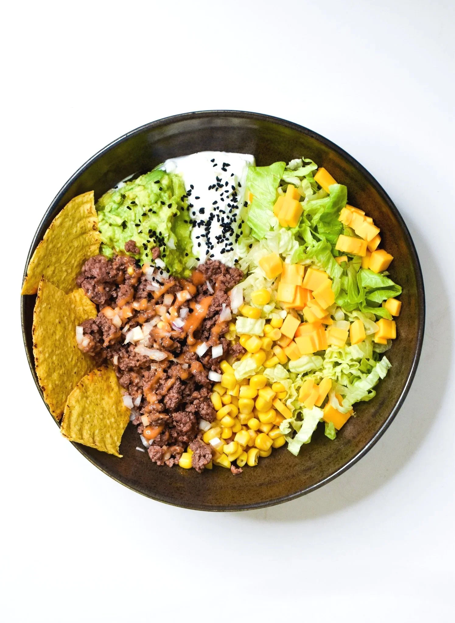 Bowl of taco salad with ground beef, shredded cheese, lettuce, diced tomatoes, sour cream, guacamole, tortilla chips, black sesame seeds, and crunchy taco shell pieces, on a white surface.