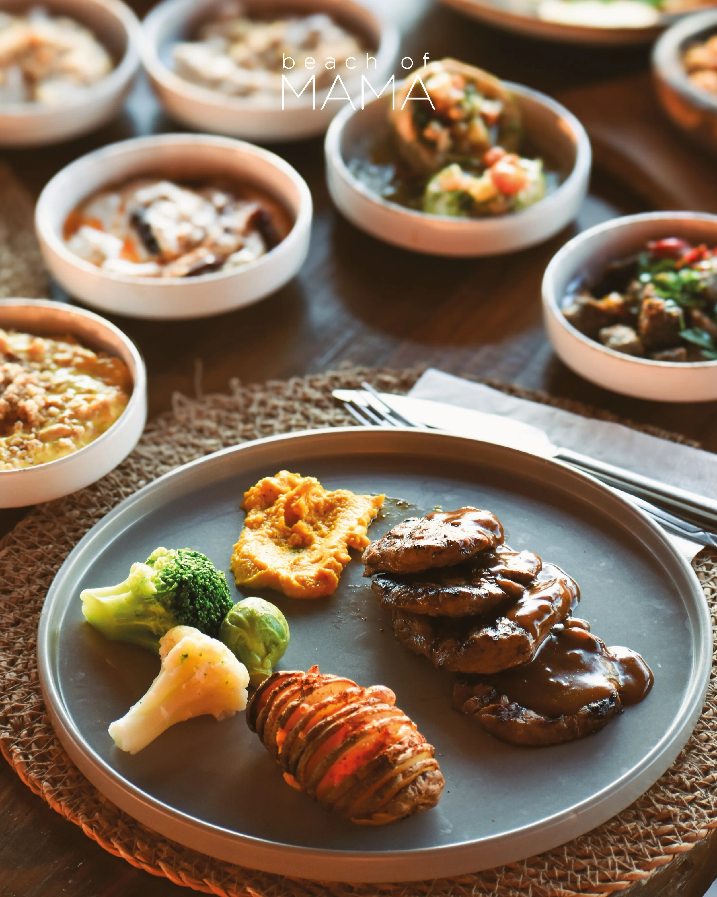 Plate with mashed sweet potatoes, broccoli, and cauliflower, along with grilled hamburger patties topped with gravy and a Hasselback potato with seasoning. Several small bowls in the background contain various dishes.
