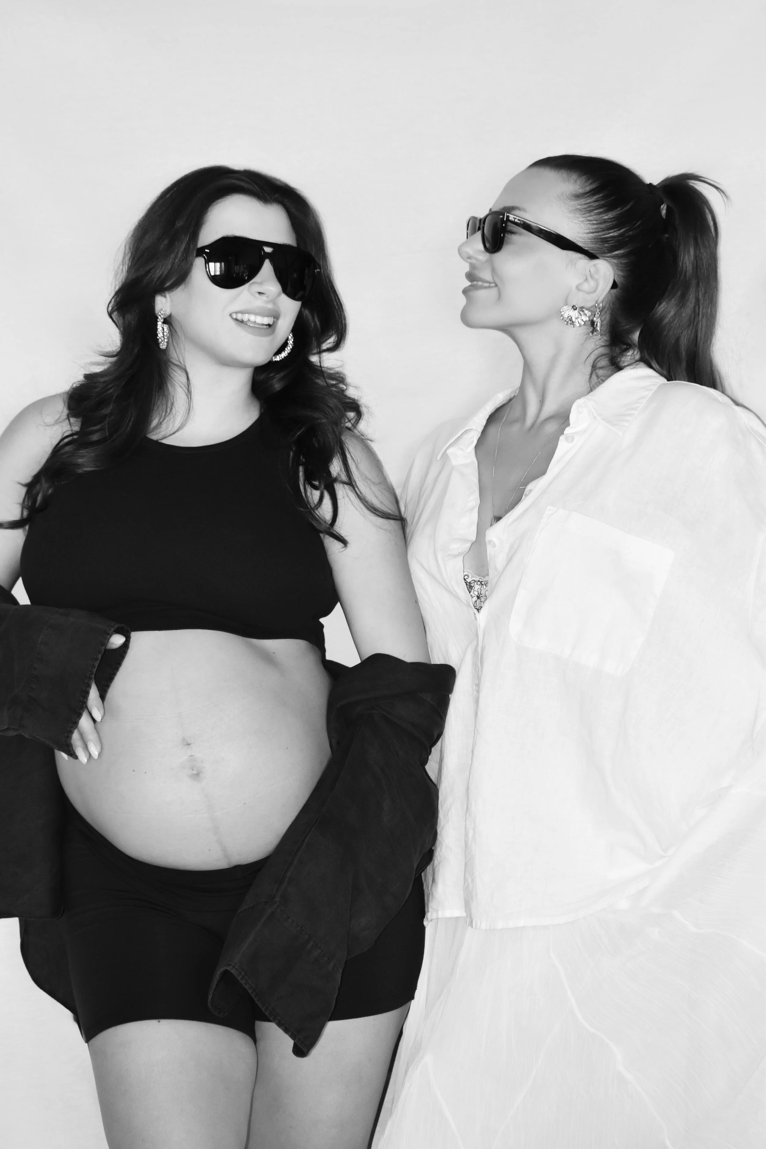 Two women wearing sunglasses, smiling, standing close to each other against a plain background. The woman on the left has long dark hair, wears a black sleeveless top, and has her jacket draped over her arm. The woman on the right has her hair tied back, wears a light-colored button-up shirt, and appears to be pregnant.