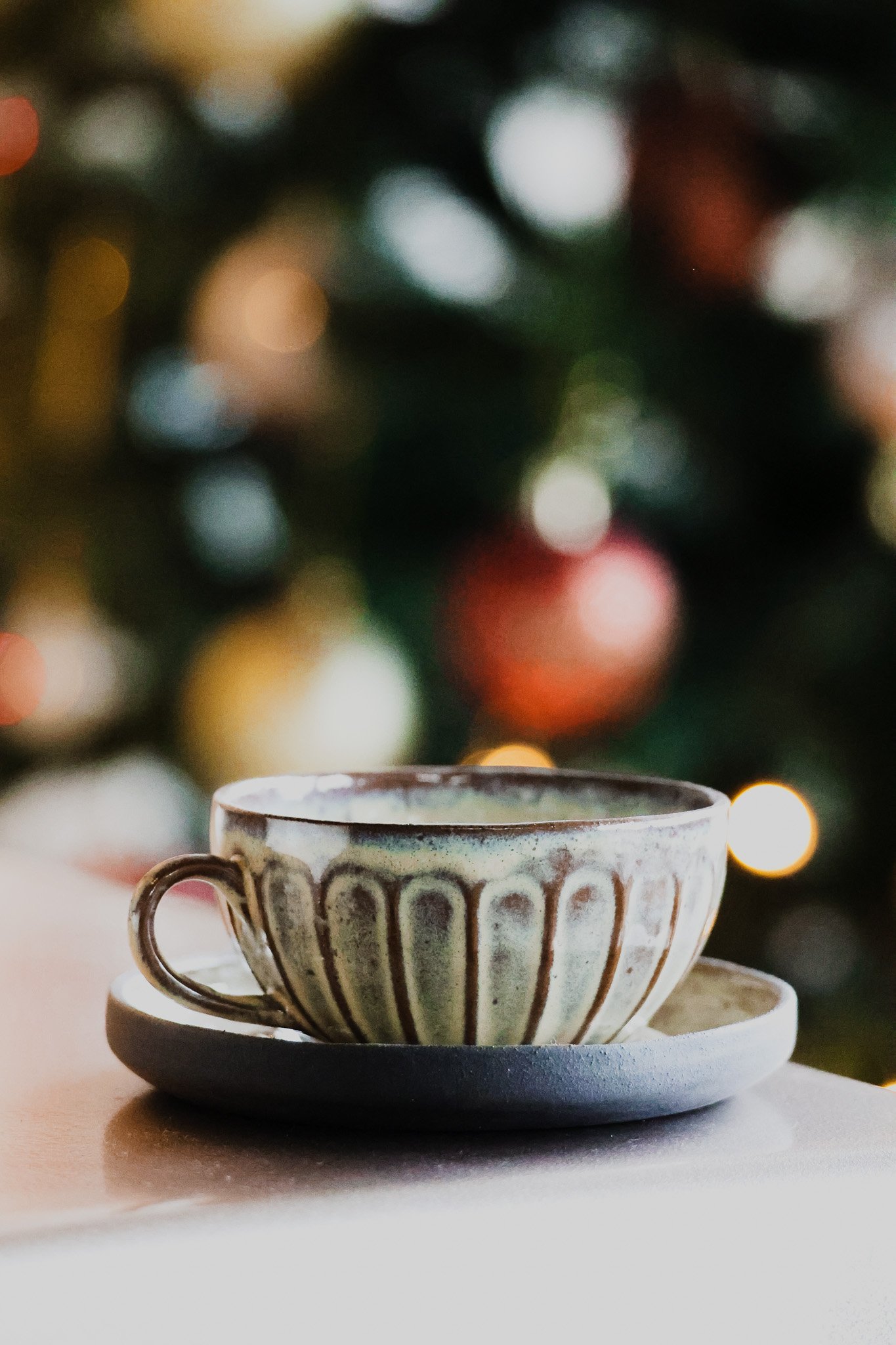 A ceramic cup with a saucer on a table, with a blurred Christmas tree with lights in the background.