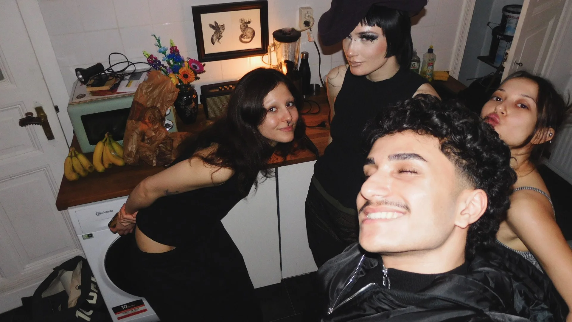 Four friends taking a selfie in a kitchen, with a woman in black with short black hair and a hat in the center, a woman with long dark hair and a septum piercing to her left, a woman with long dark hair and earrings to her right, and a man with curly hair and a black jacket smiling in front.