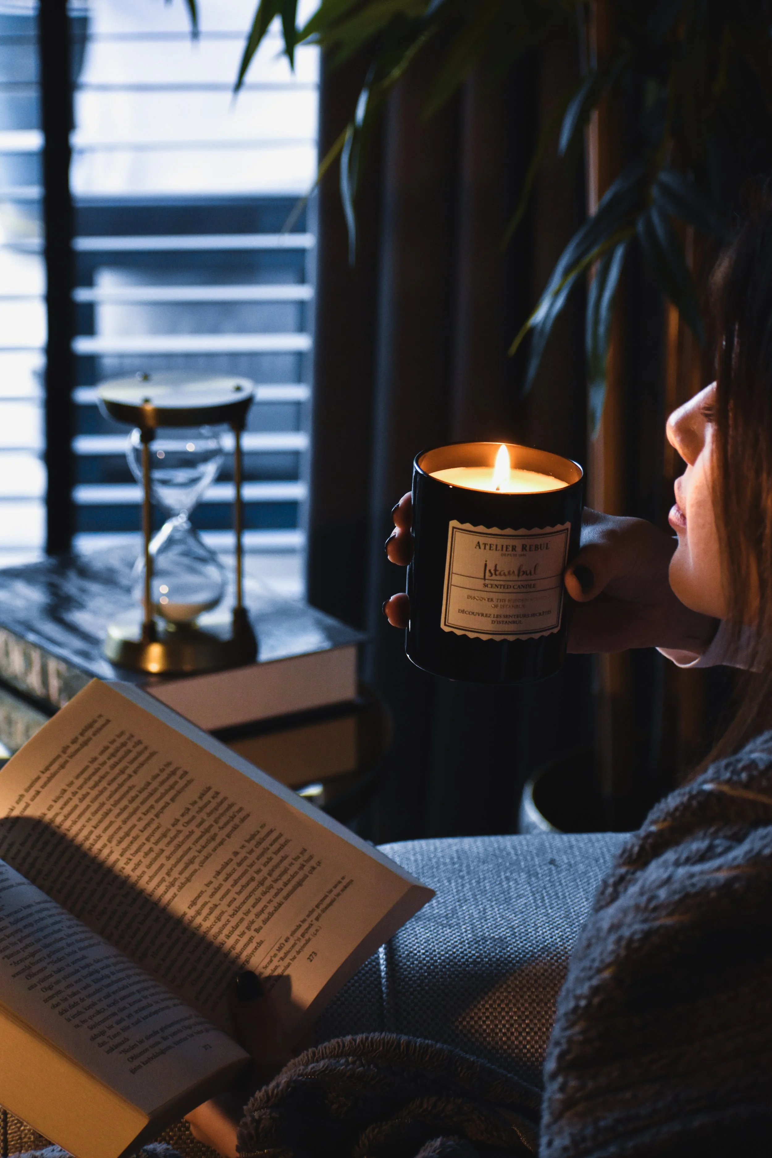 A woman sitting indoors holding a lit candle in a black jar, reading a book with warm lighting, near a window with blinds and an hourglass on a table.