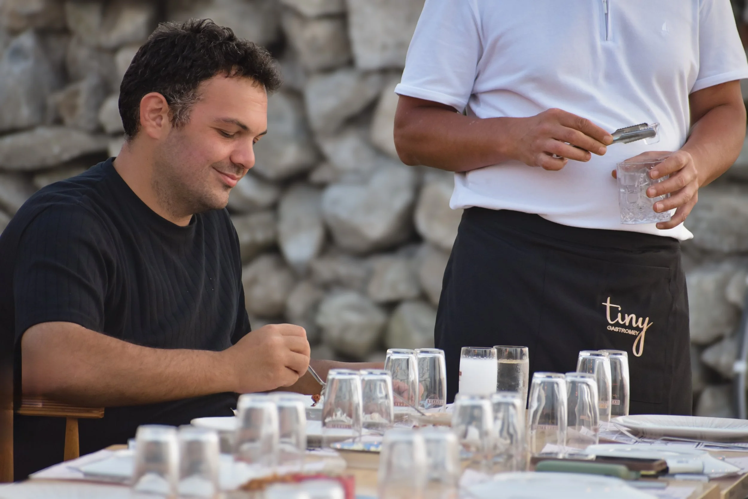 A man is seated at a table set for dining outdoors, while a server stands nearby holding a glass and a utensil.