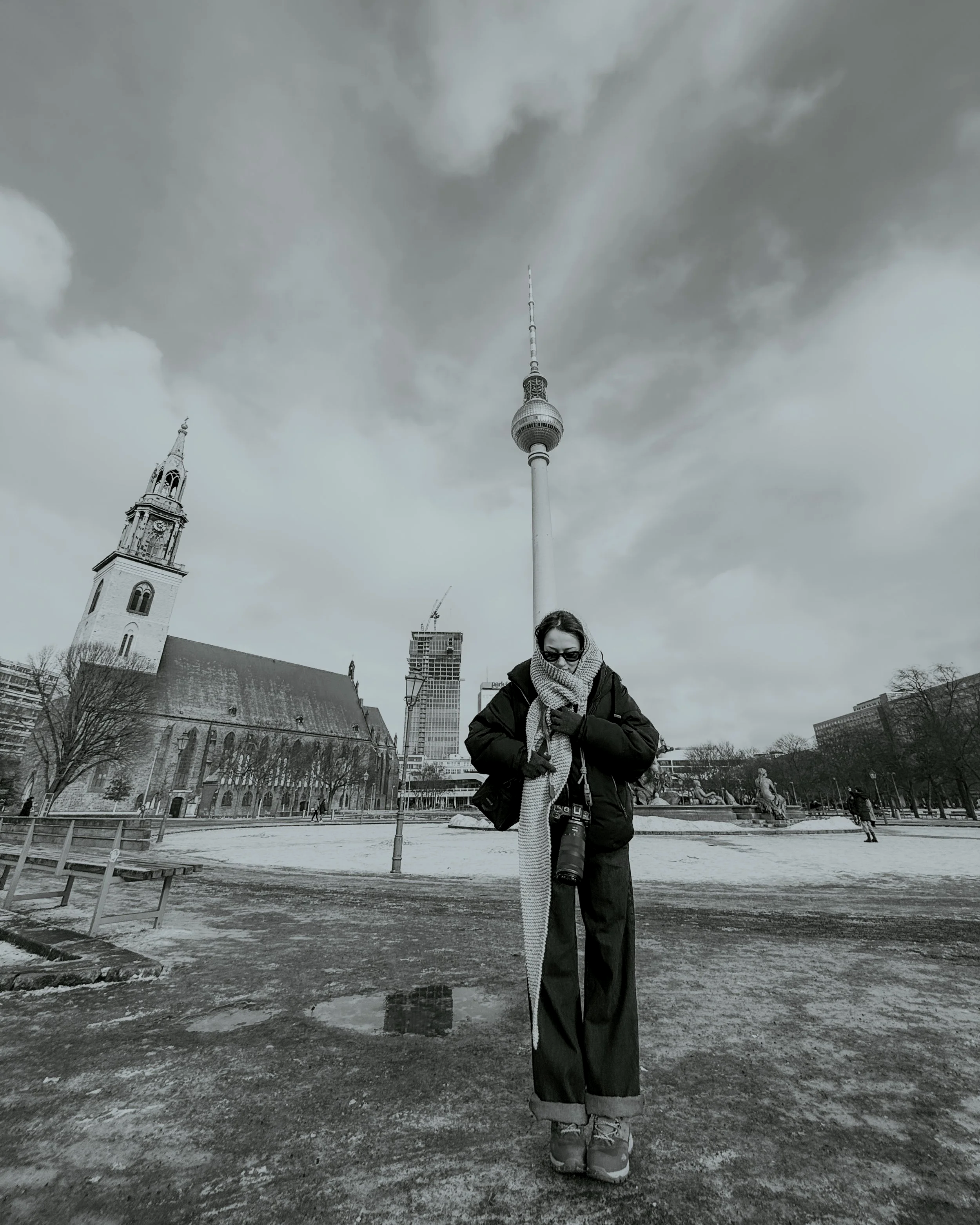 A person standing outdoors in a city with a camera around their neck, wearing a coat, scarf, and sunglasses, with a church and a tall telecommunications tower in the background. It is a winter day with snow and puddles on the ground.