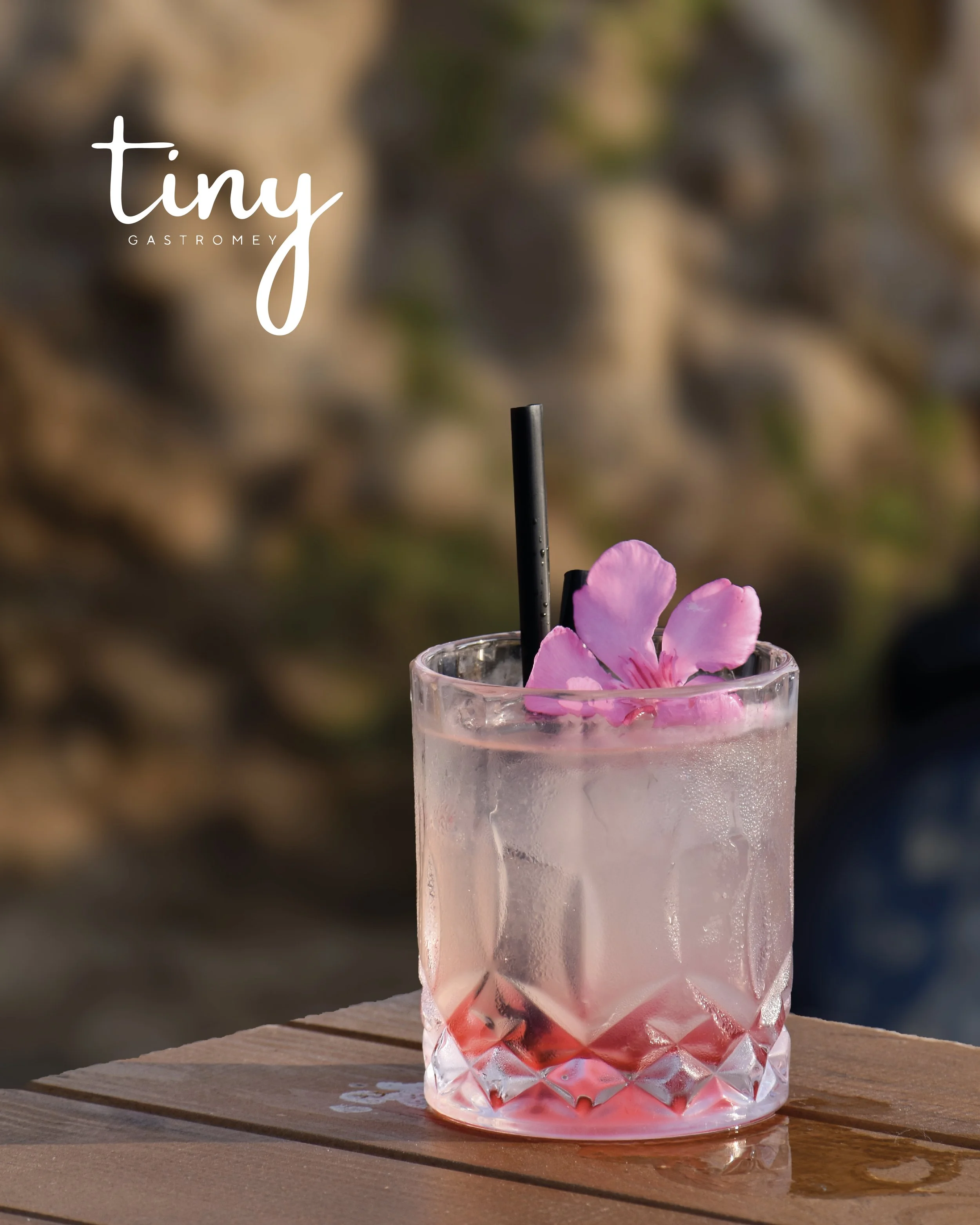A pink cocktail in a textured glass with a black straw and pink flower garnish on a wooden surface outdoors.