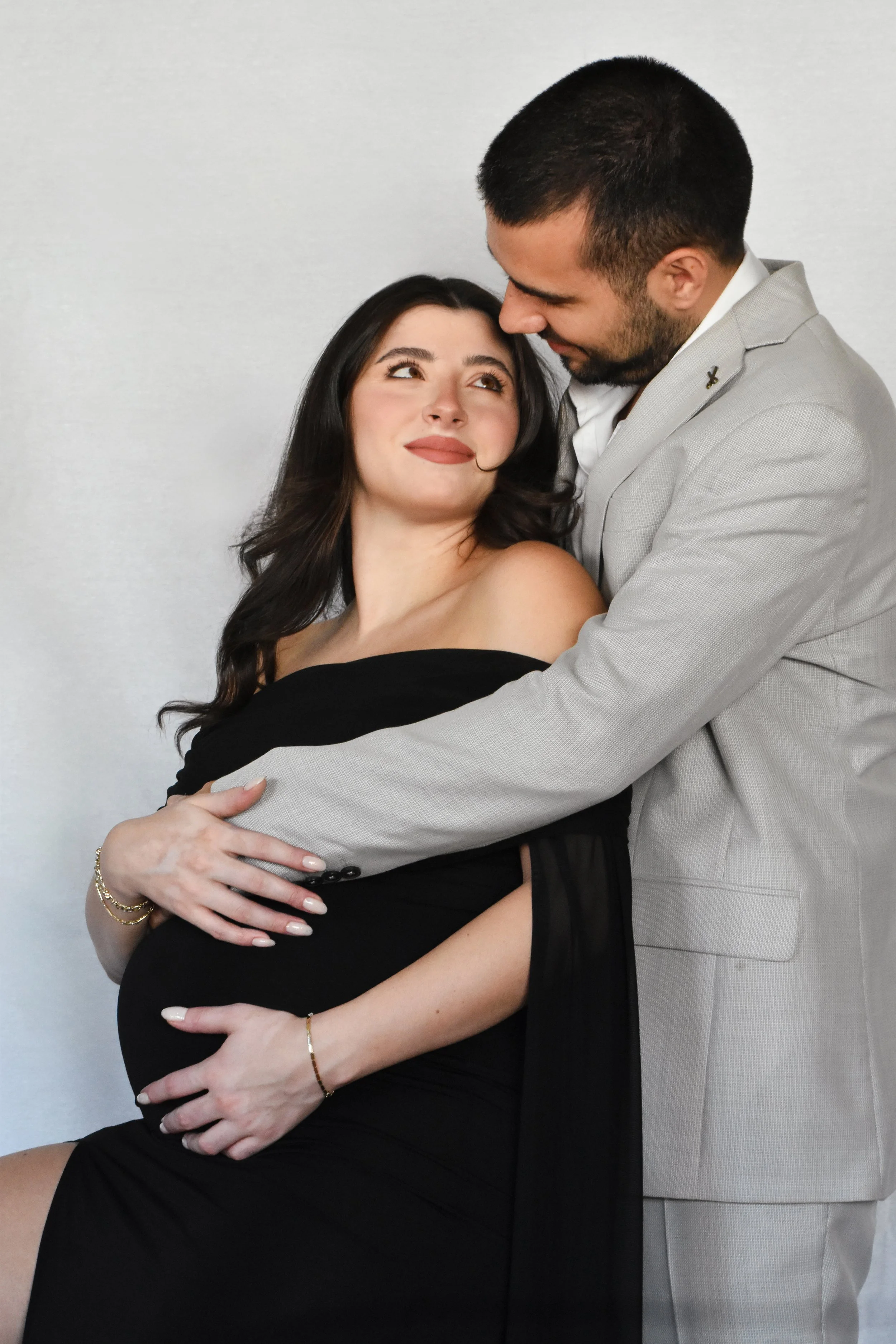 A pregnant woman with long dark hair and makeup, sitting down in a black off-shoulder dress, being embraced by a man in a light gray suit, both smiling and looking at each other affectionately against a plain white background.