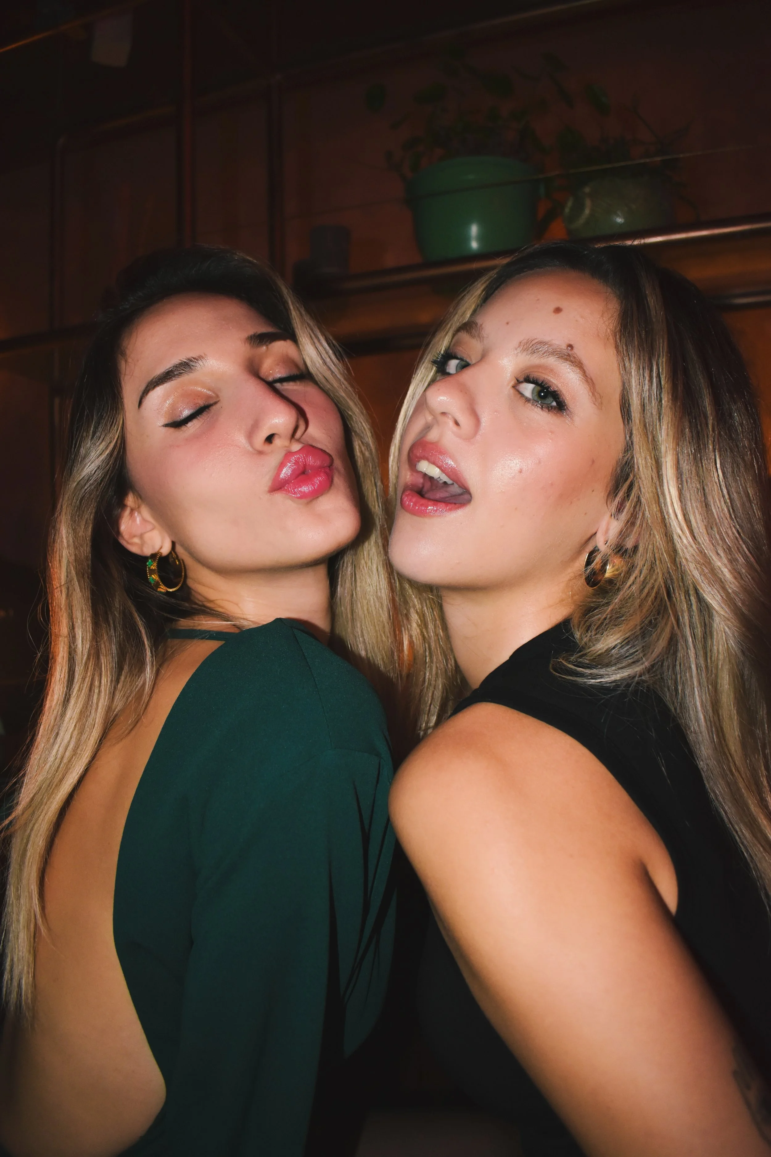 Two young women with long hair posing closely, making playful facial expressions in a dimly lit indoor setting.
