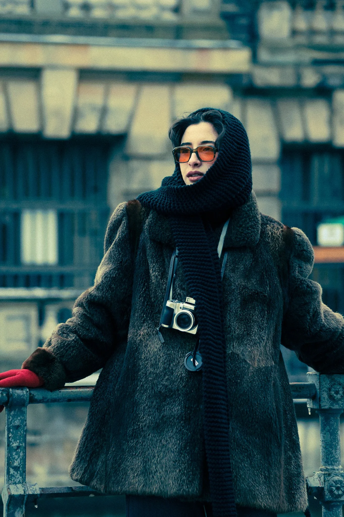 A woman wearing large black scarf, sunglasses, a fur coat, red gloves, and a camera around her neck stands outdoors near a railing with an urban background.