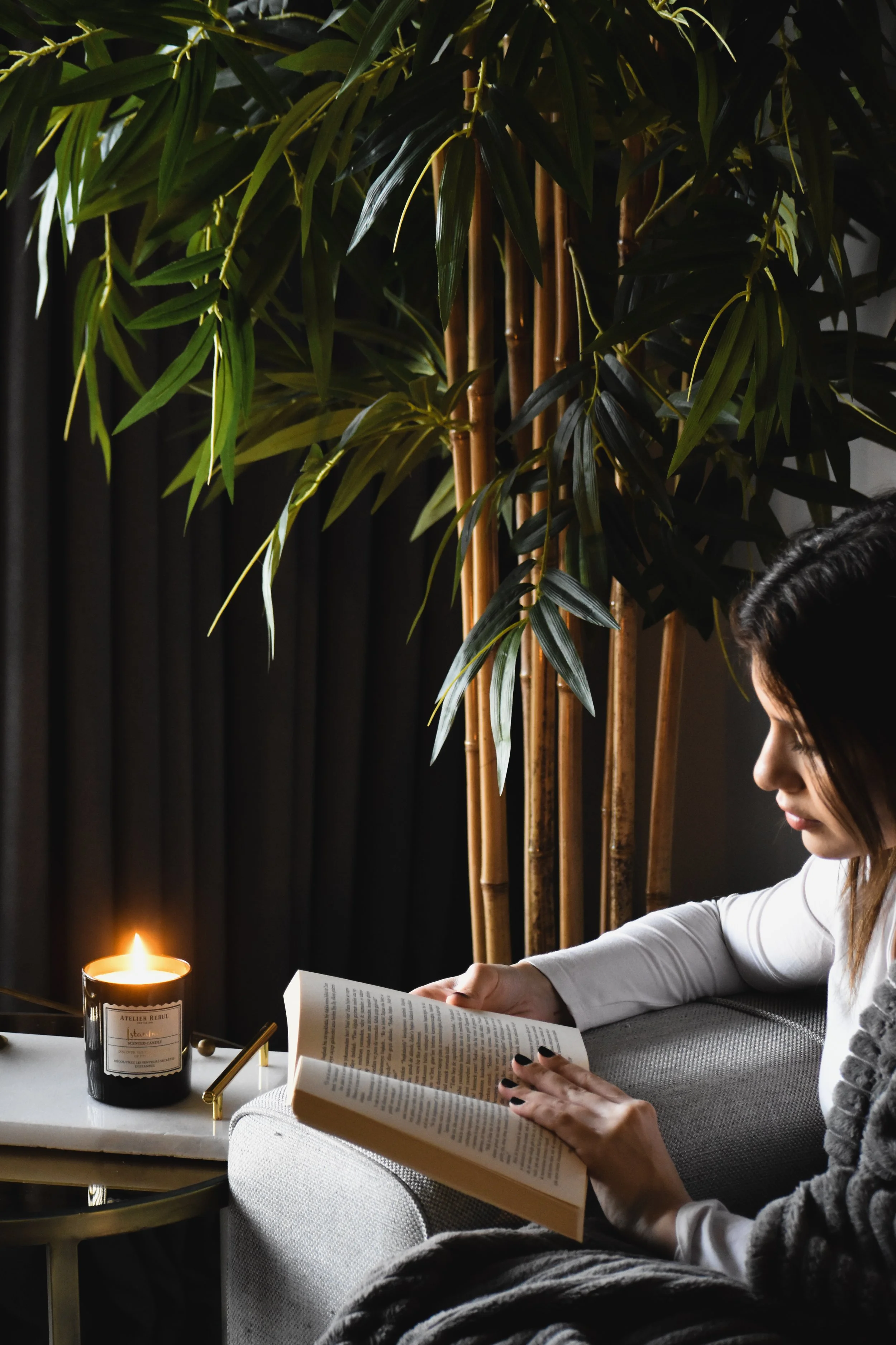 A woman with black hair reading a book on a gray couch, with a lit candle on a side table nearby, in a room with large green plants and black curtains.