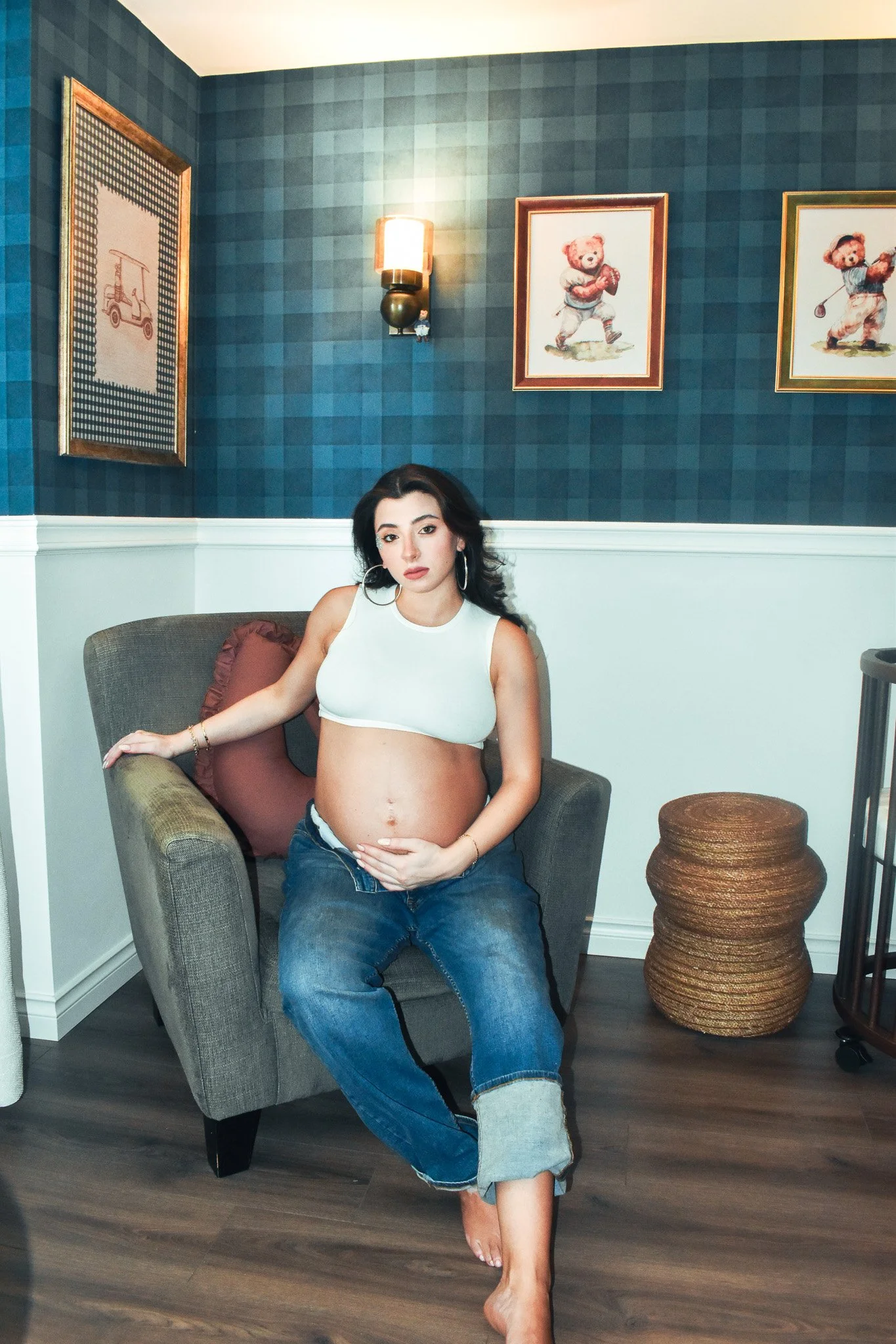 A pregnant woman with long dark hair, wearing hoop earrings, a white sleeveless crop top, and loose blue jeans, sitting barefoot on a brown armchair in a room with blue checkered wallpaper and framed teddy bear pictures, resting her hand on her belly and the armrest.