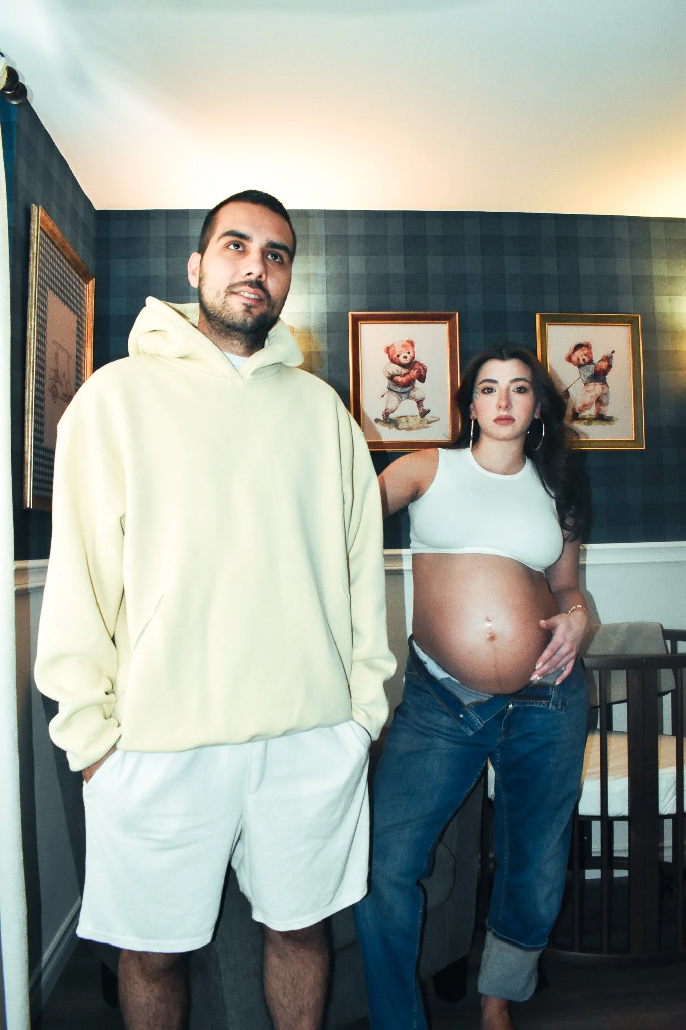 A man and a pregnant woman standing indoors in front of framed teddy bear artwork on a wall.