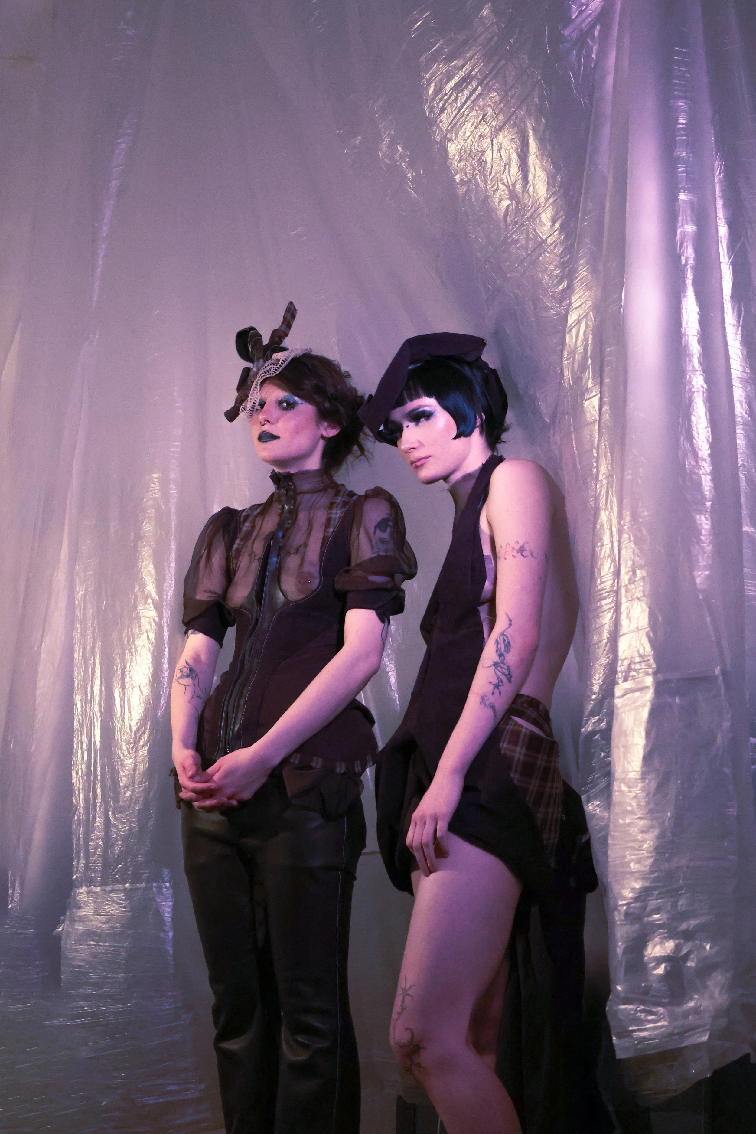Two women with bold makeup and tattoos pose against a purple metallic backdrop, wearing dark, avant-garde fashion with hats.