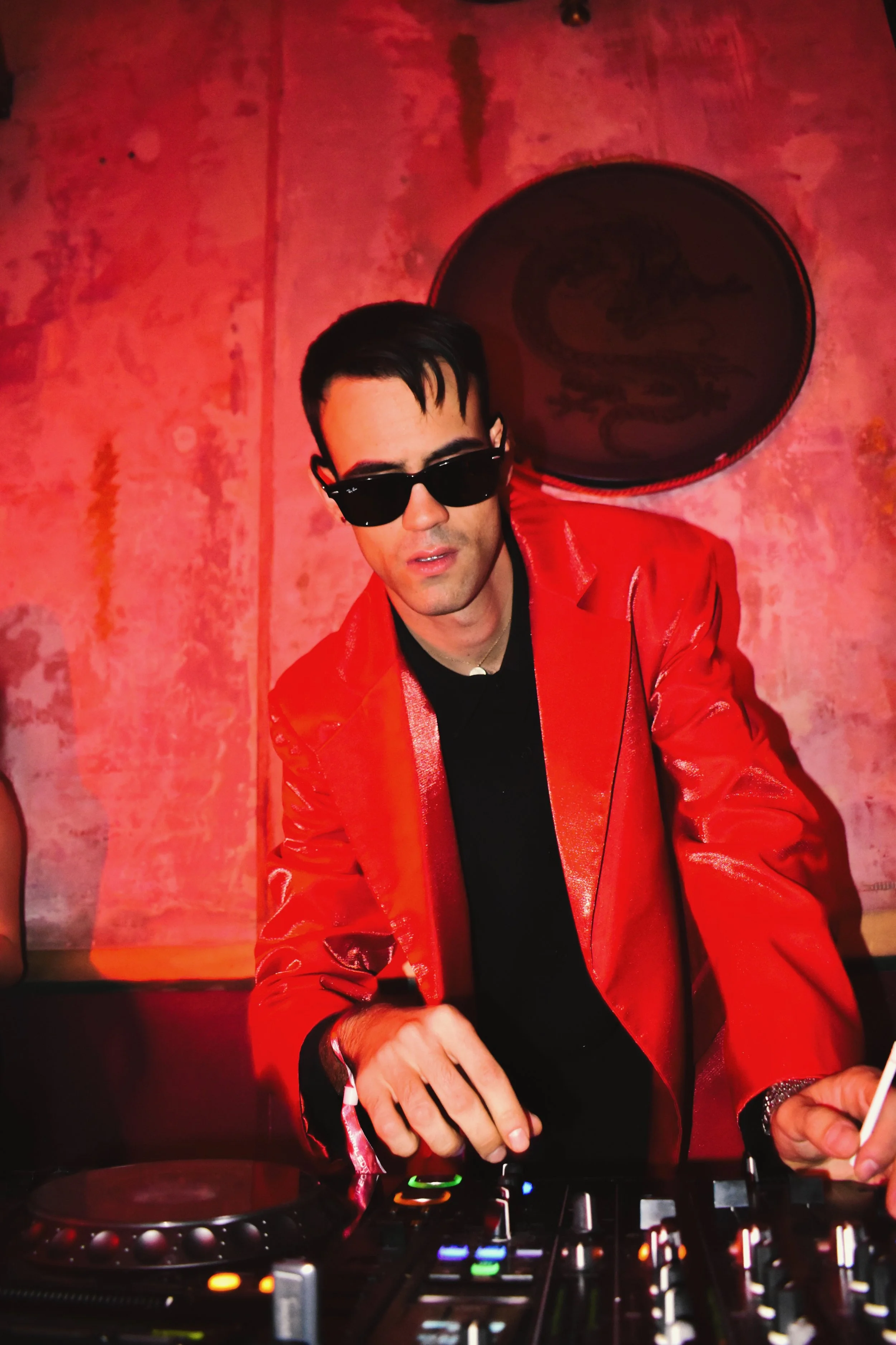 A DJ wearing a red leather jacket and black sunglasses is operating a DJ mixer under red lighting.
