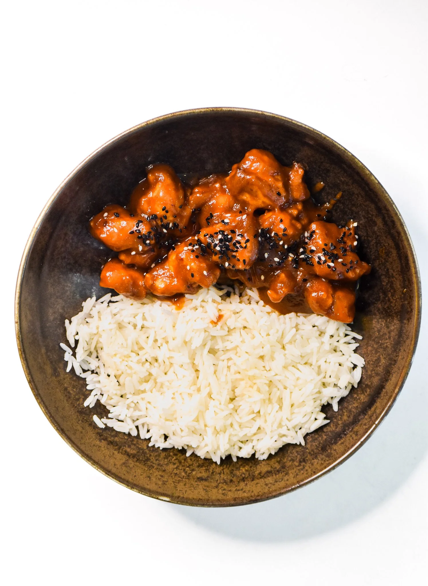 A bowl with white rice and sweet and sour chicken topped with black sesame seeds.