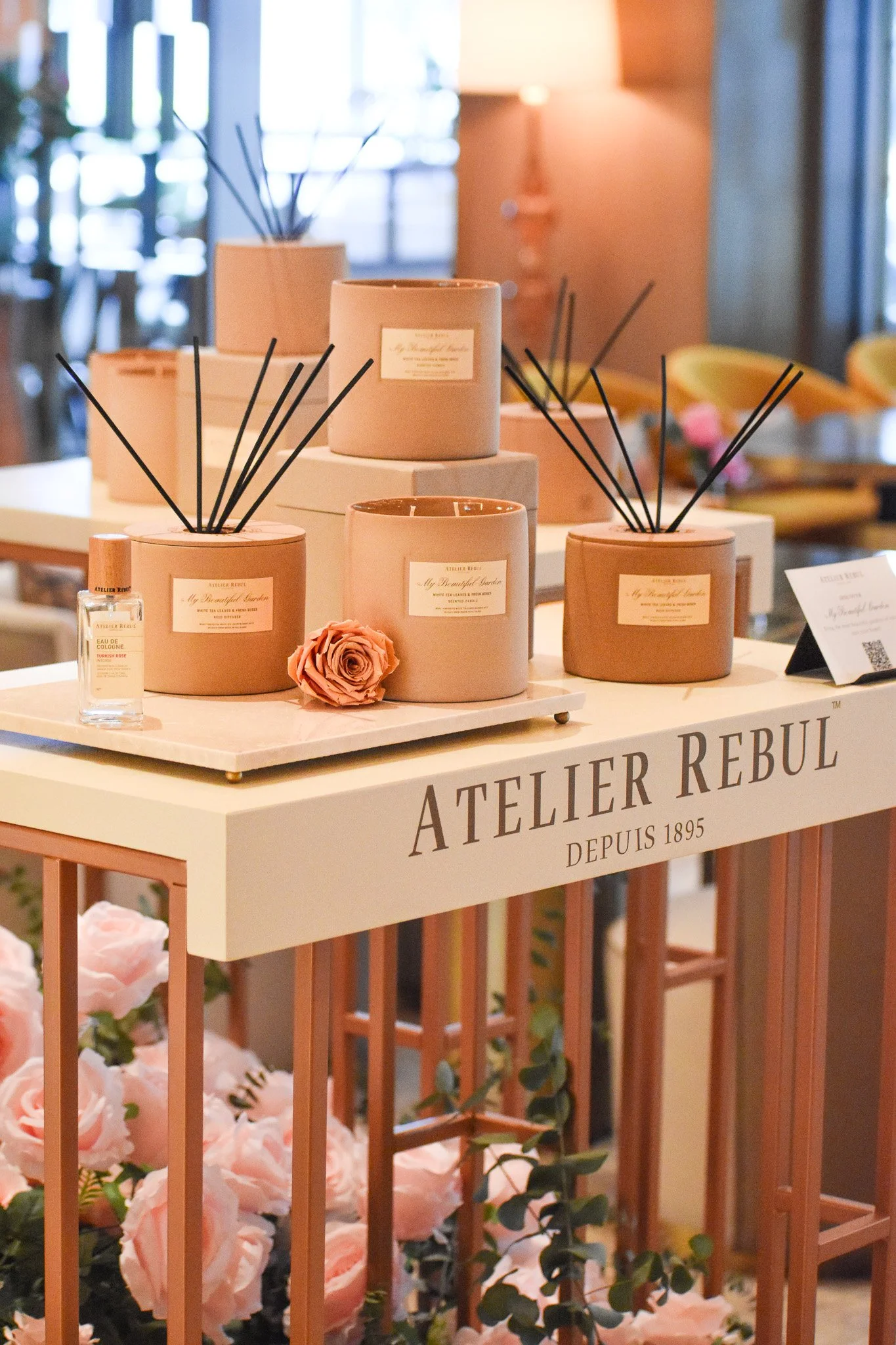 Display of scented candles and a small perfume bottle on a white table with pink roses underneath, at an event for Atelier Rebul, decorated in soft pink tones.