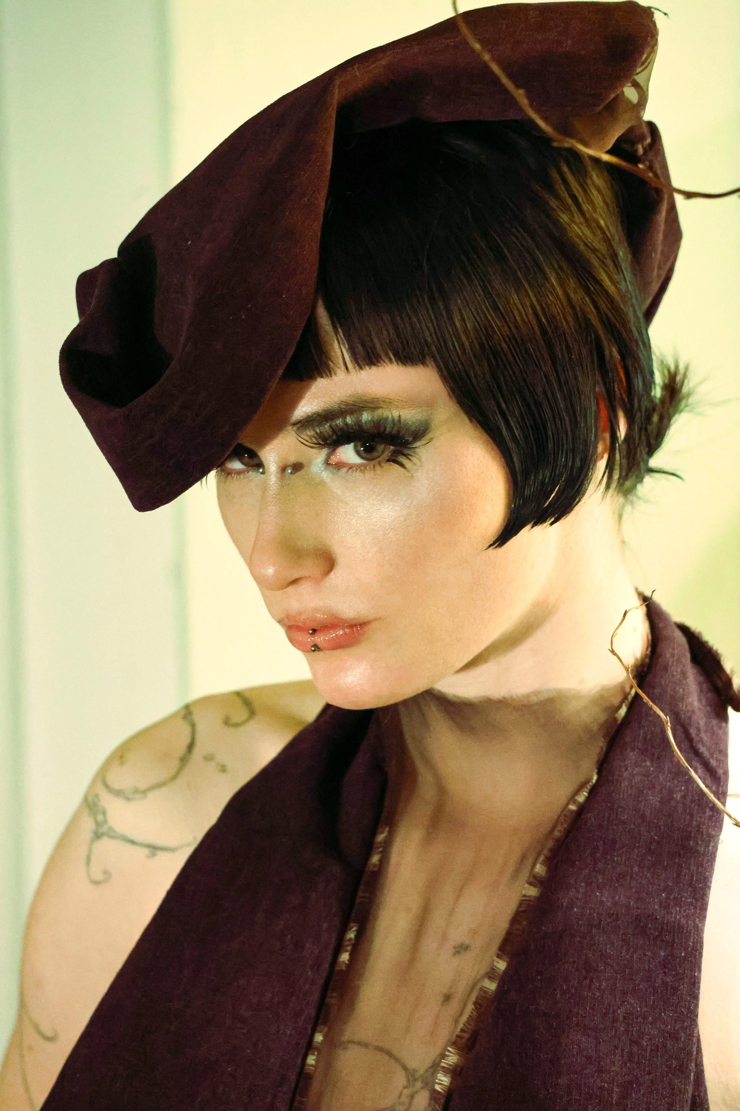A woman with short dark hair, green eye makeup, and a piercing above her lip, wearing a maroon hat and matching garment.