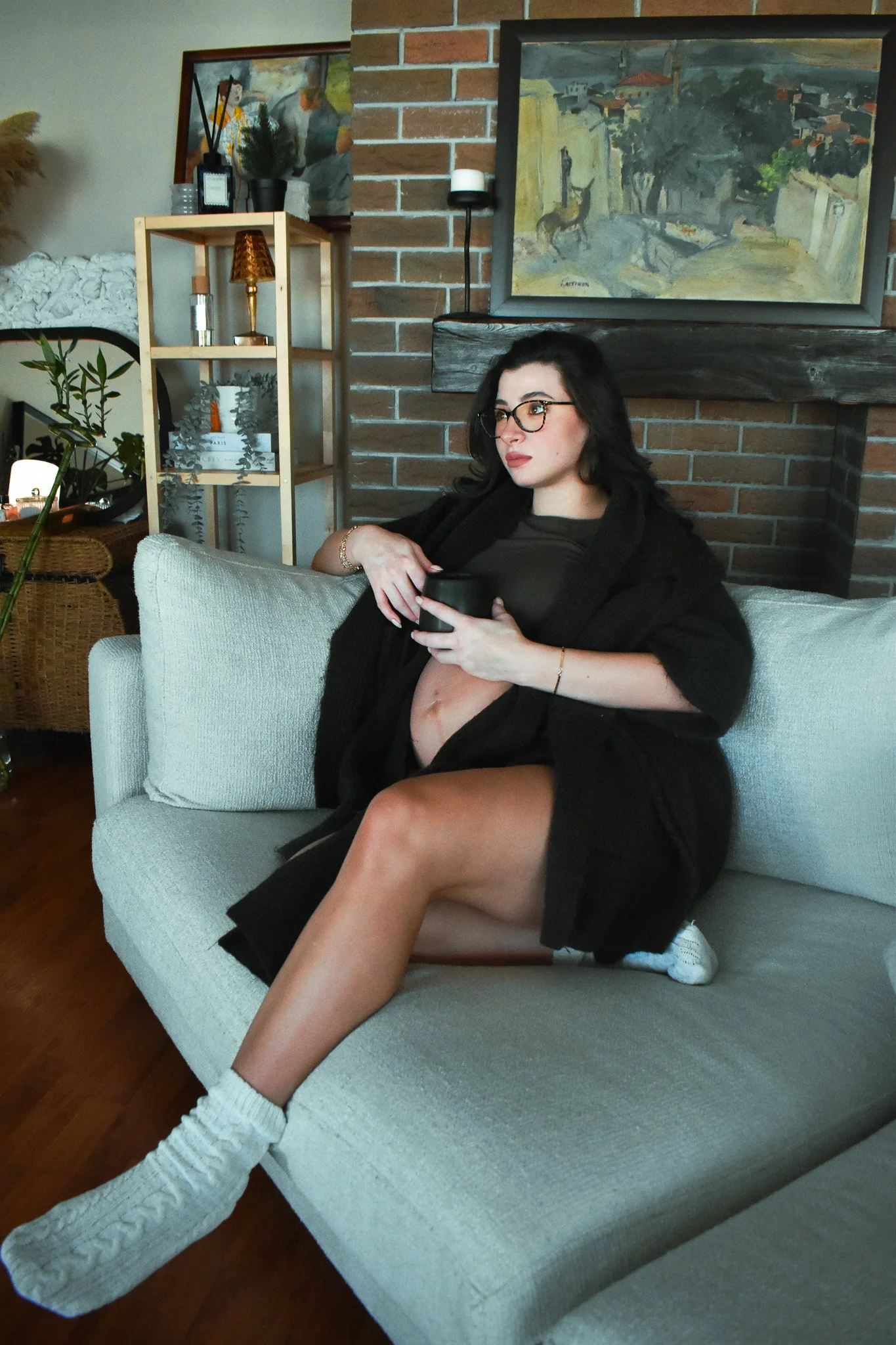 A woman with black hair and glasses sitting on a sofa, holding a phone, with her legs partially exposed, in a cozy, decorated living room.