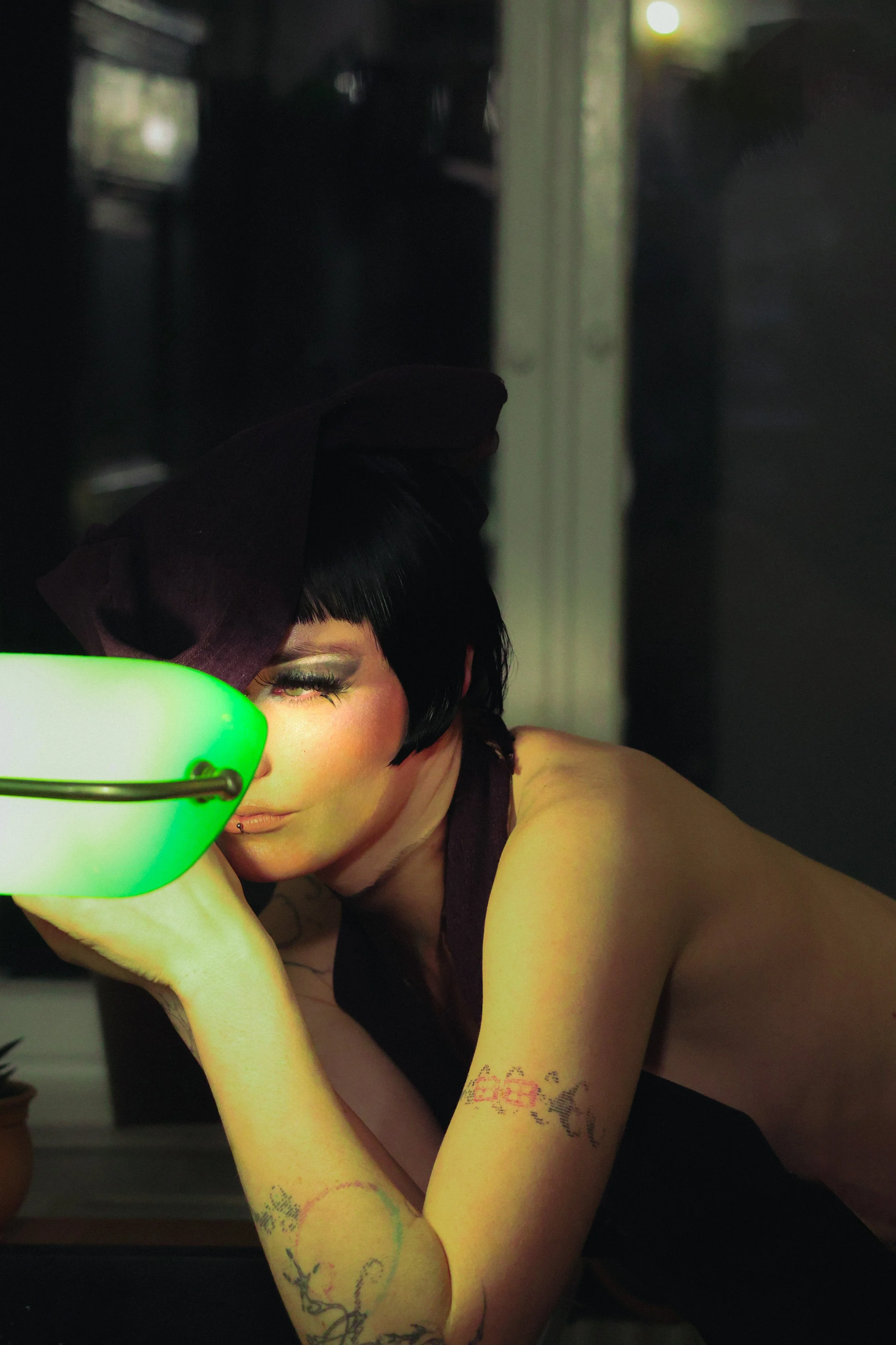 A person with short black hair, makeup, and tattoos on their arms, resting their head on their hand, illuminated by a glowing green light, in a dark room.