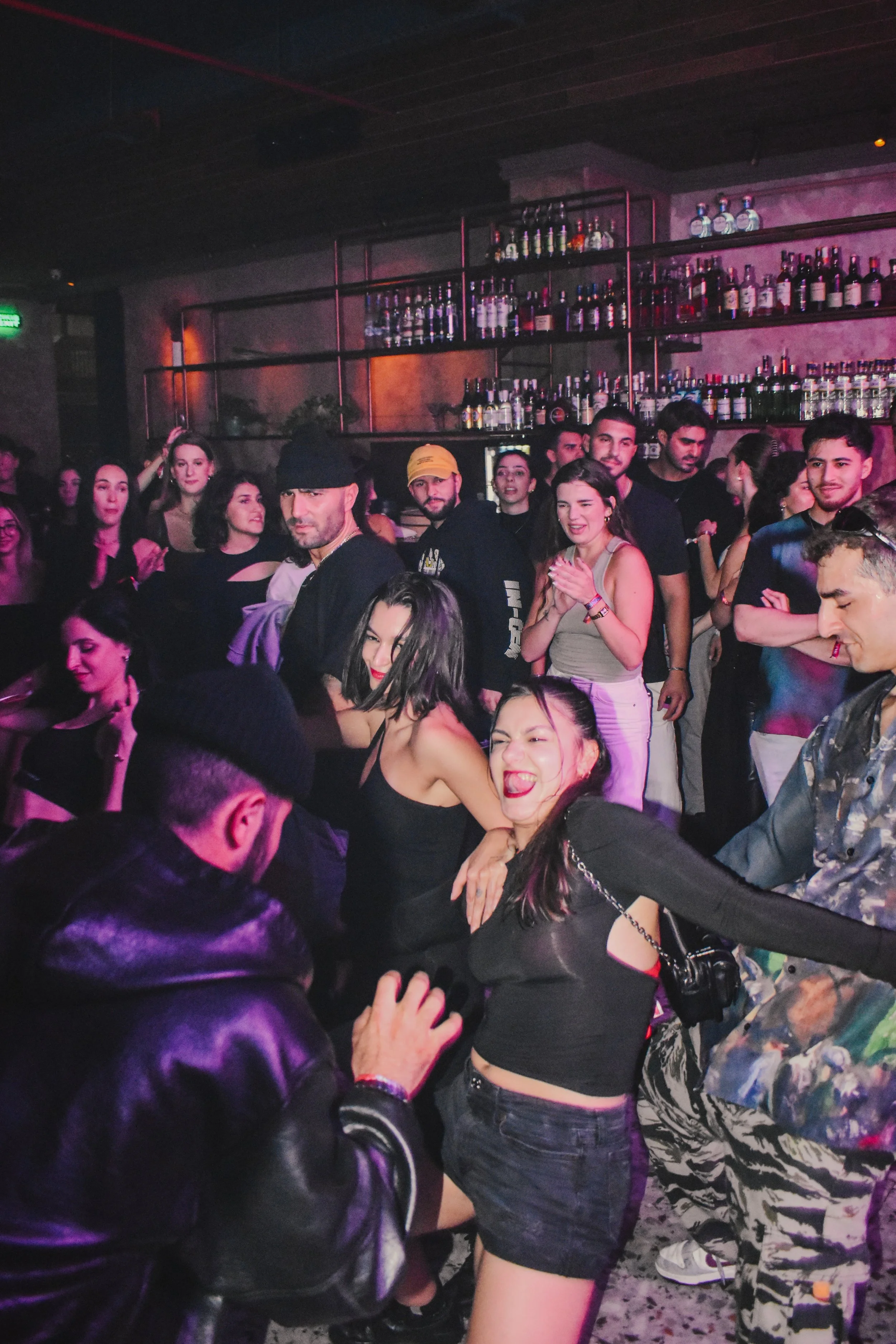 People dancing and socializing in a dark nightclub with shelves of liquor bottles on the wall in the background.