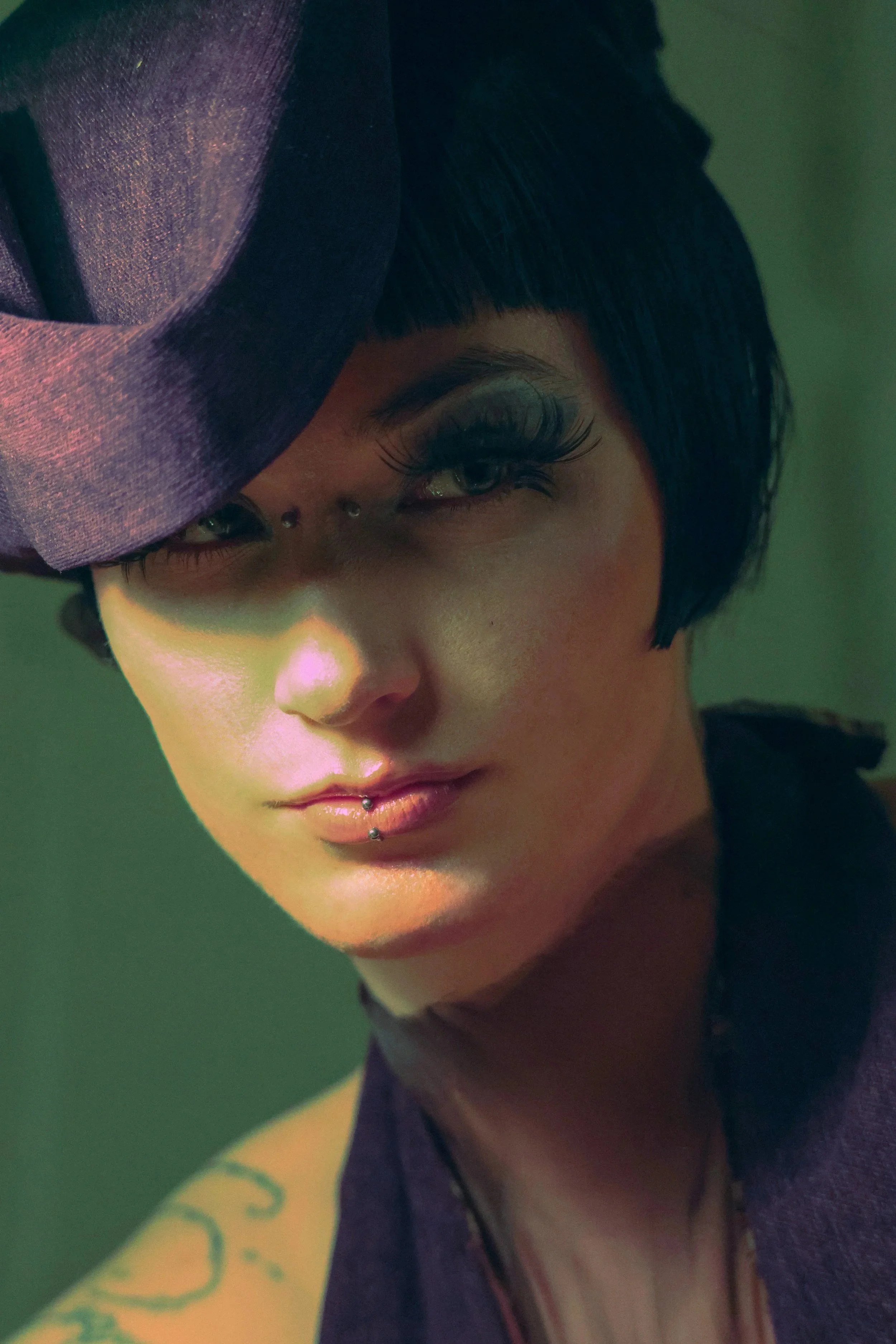A person with short black hair, makeup, and a new lip piercing, posing against a green background, wearing a purple garment and holding a purple hat near their face.