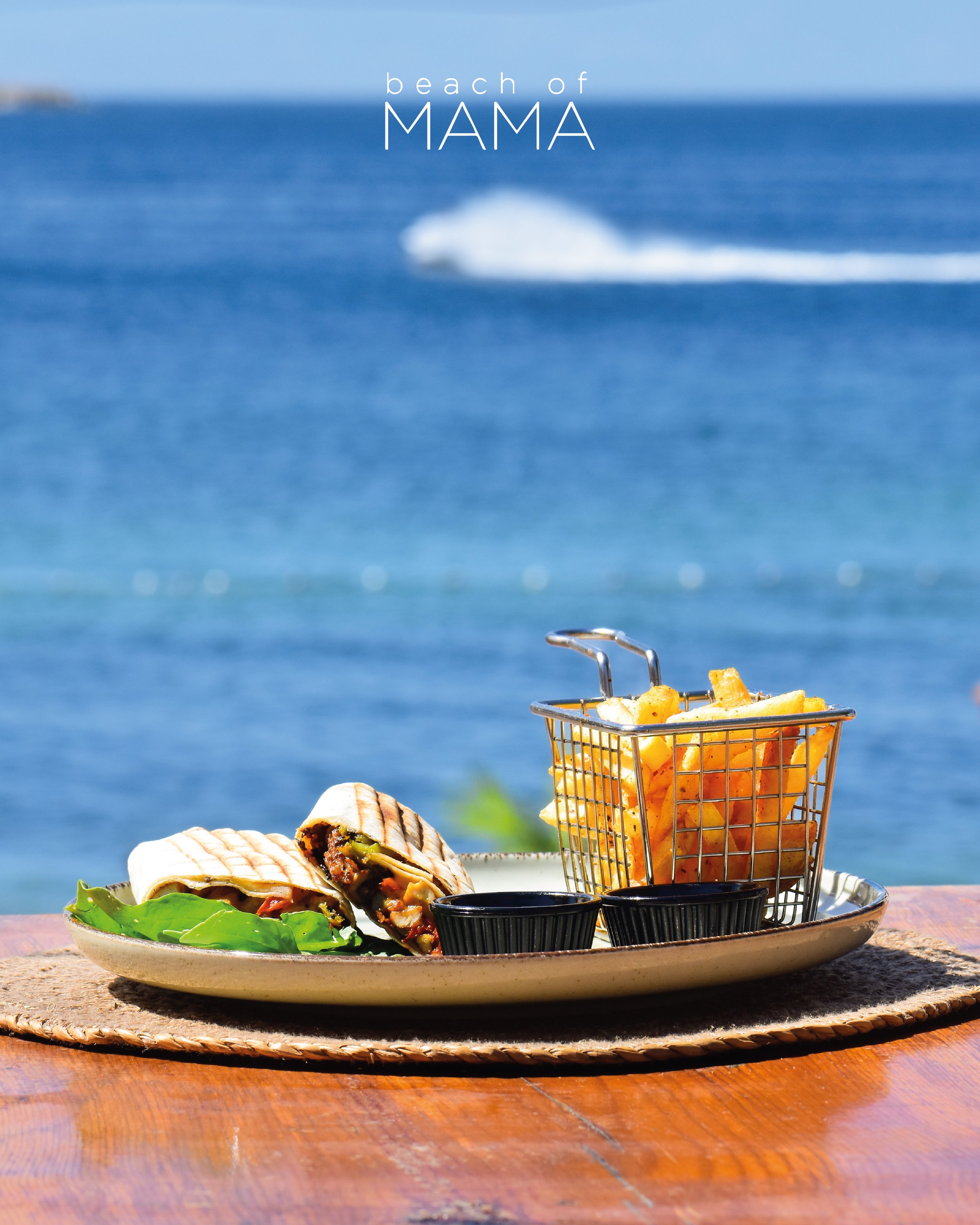 A plate with a grilled wrap sandwich, a basket of French fries, and two small containers of dipping sauce on a wooden table by the ocean, with a boat in the background.
