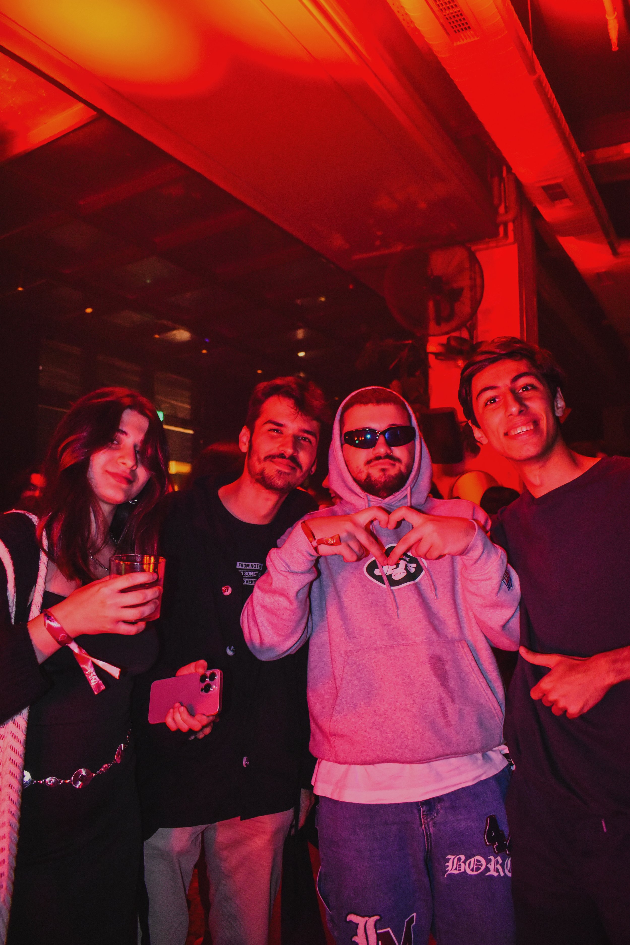 Four young adults at a nightclub, with dim red lighting, standing close together, smiling, and posing for a photo, one holding a drink and another making a heart shape with their hands.