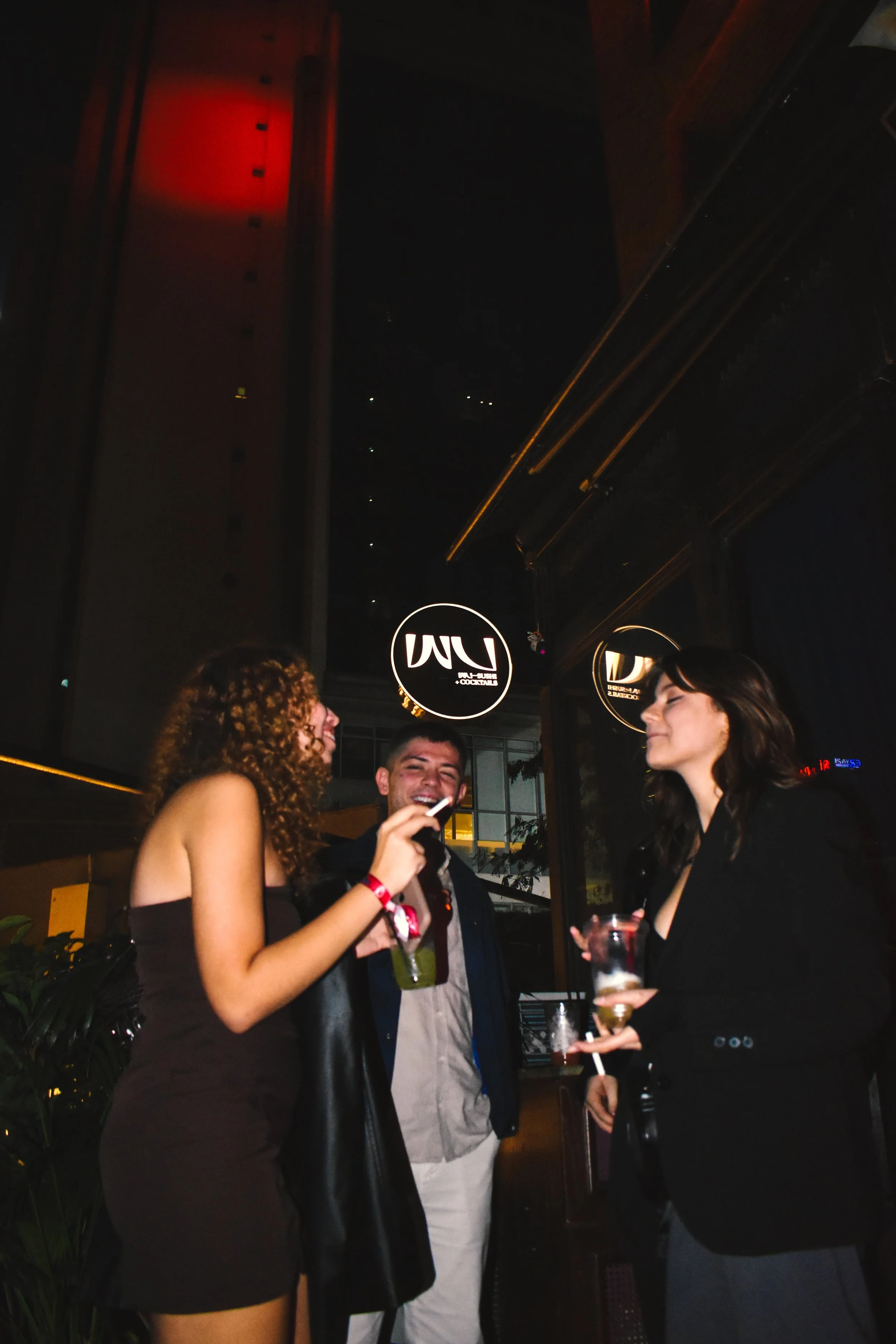 Three friends standing inside at night, smiling and chatting, holding drinks, with a hanging round sign that reads 'LUC' in the background.