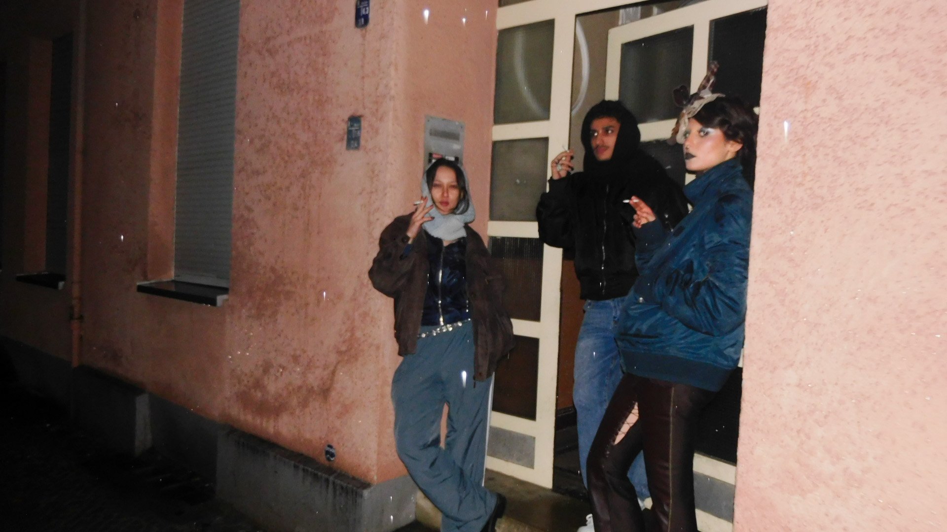 Three young people standing outside at night, leaning against a pinkish stucco wall near a door. One person is smoking, another appears to be making a pouty face, and the third has long dark hair and dark lipstick, dressed in dark clothing.