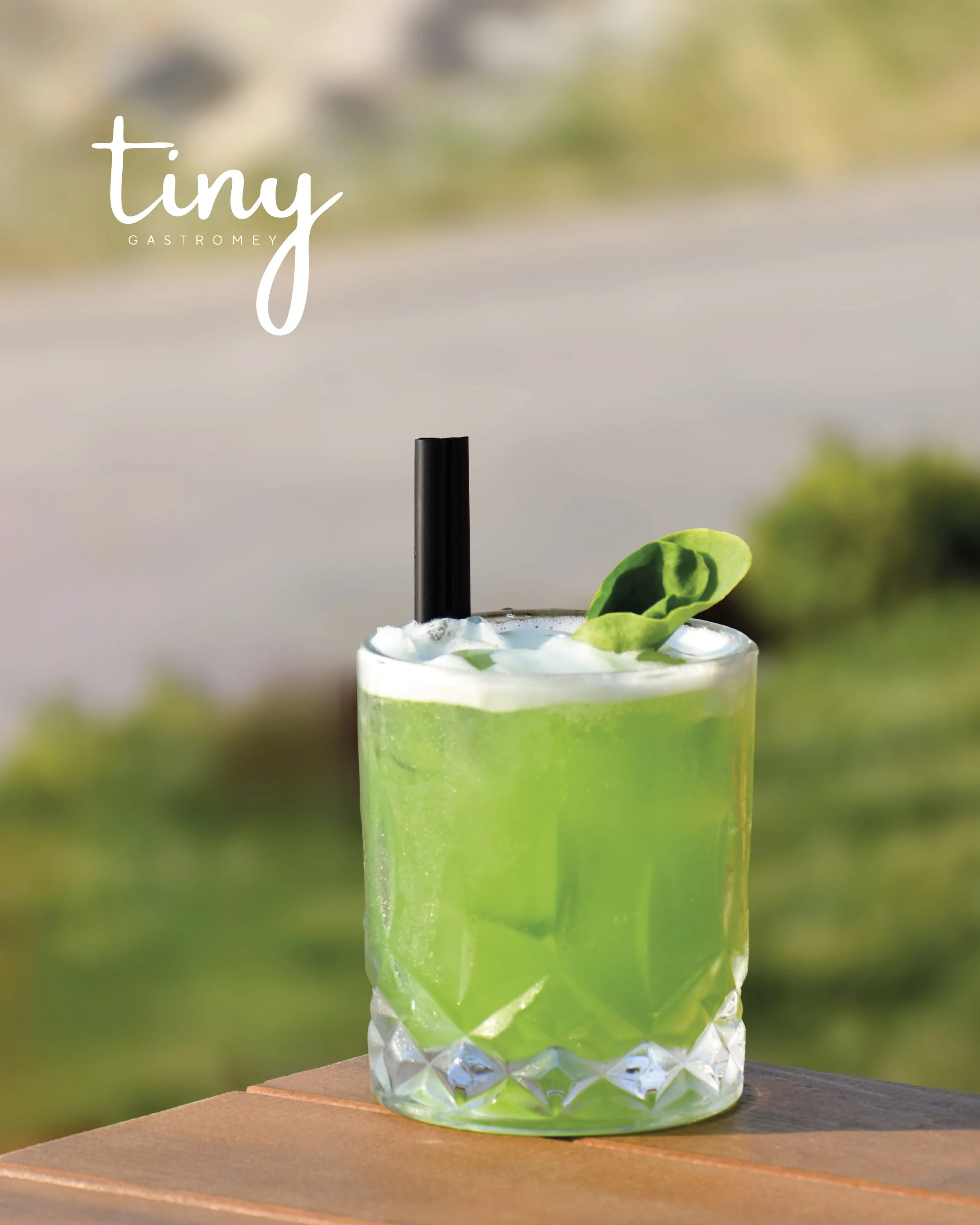 A bright green cocktail in a textured, clear glass with a black straw and a basil leaf garnish, placed on a wooden surface outdoors.