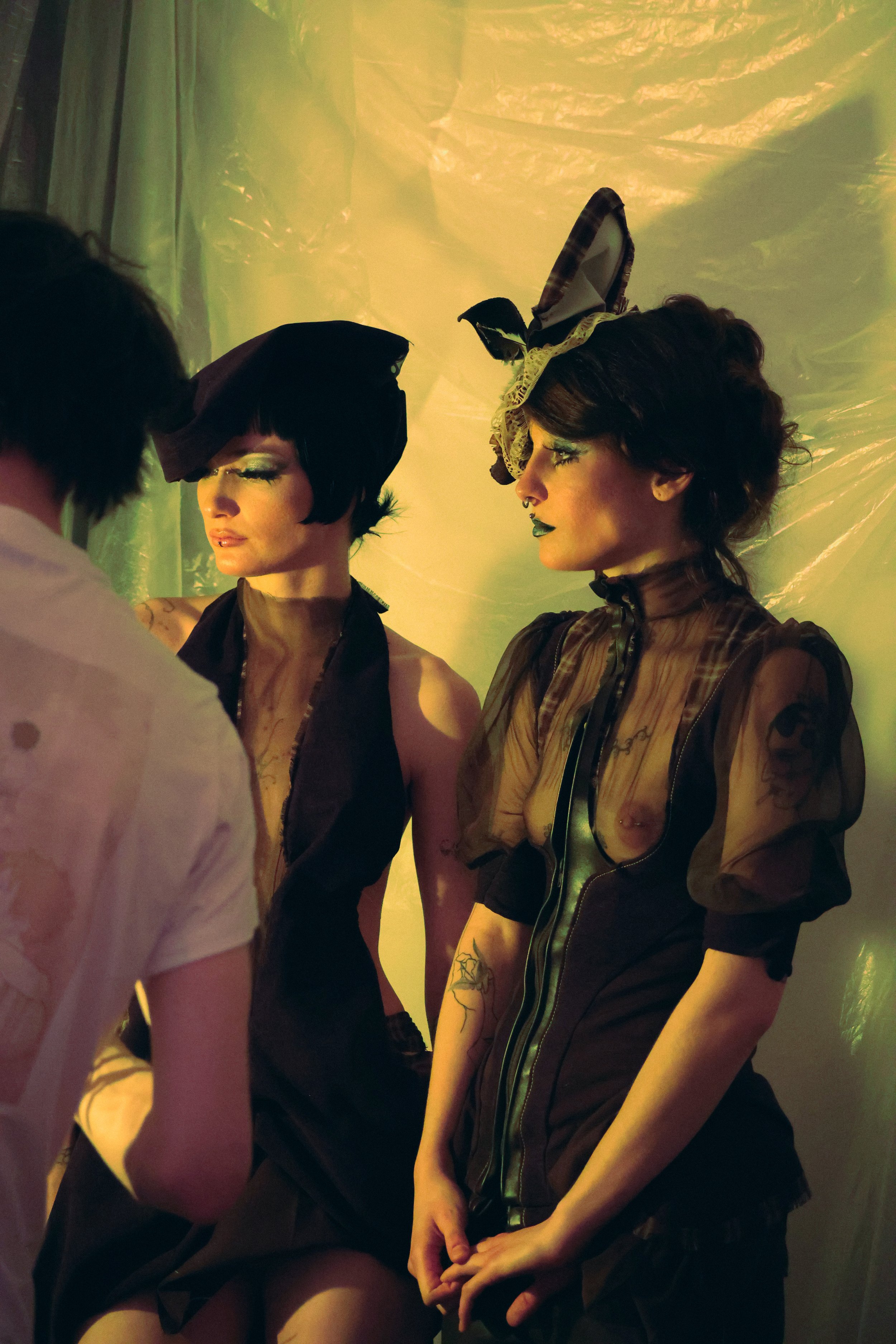 Two women dressed in elaborate goth or steampunk-style costumes with bold makeup and tattoos, standing indoors against a yellowish-green backdrop, engaging with a person whose back is to the camera.