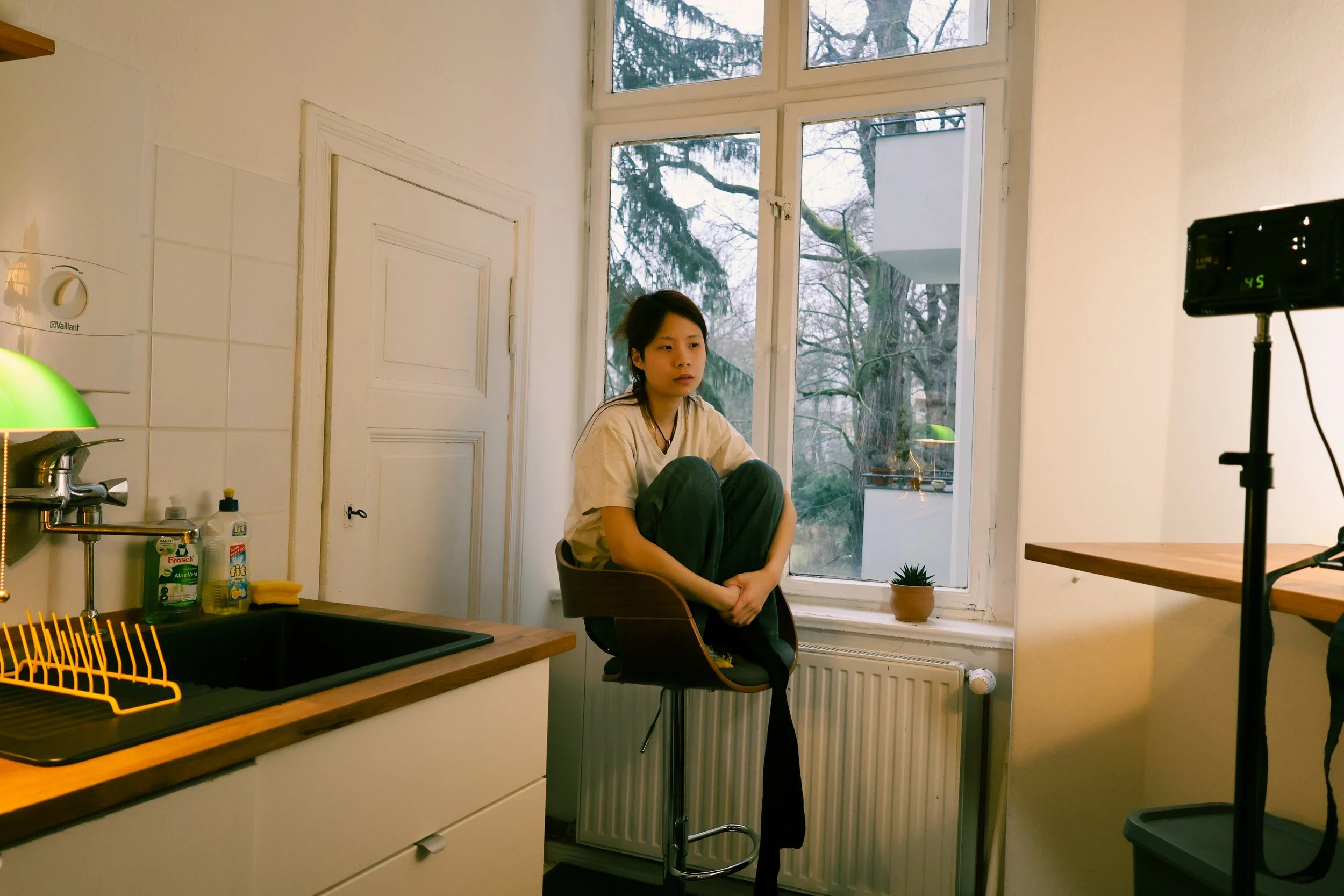 Young woman sitting on a high chair in a kitchen near a large window, looking contemplative.