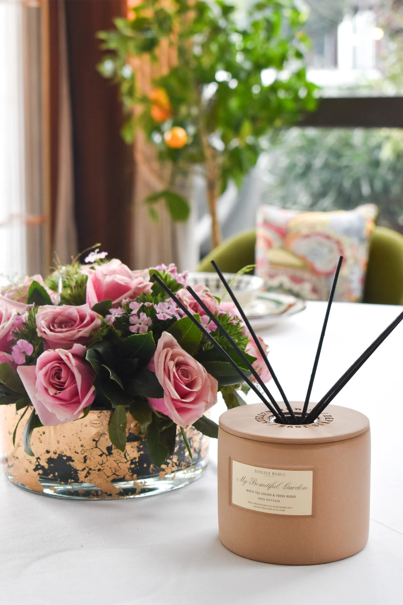 A flower arrangement of pink roses and greenery in a clear glass vase, placed on a white tablecloth next to a beige reed diffuser container with black reed sticks. In the background, a green chair and a window with greenery outside are visible.