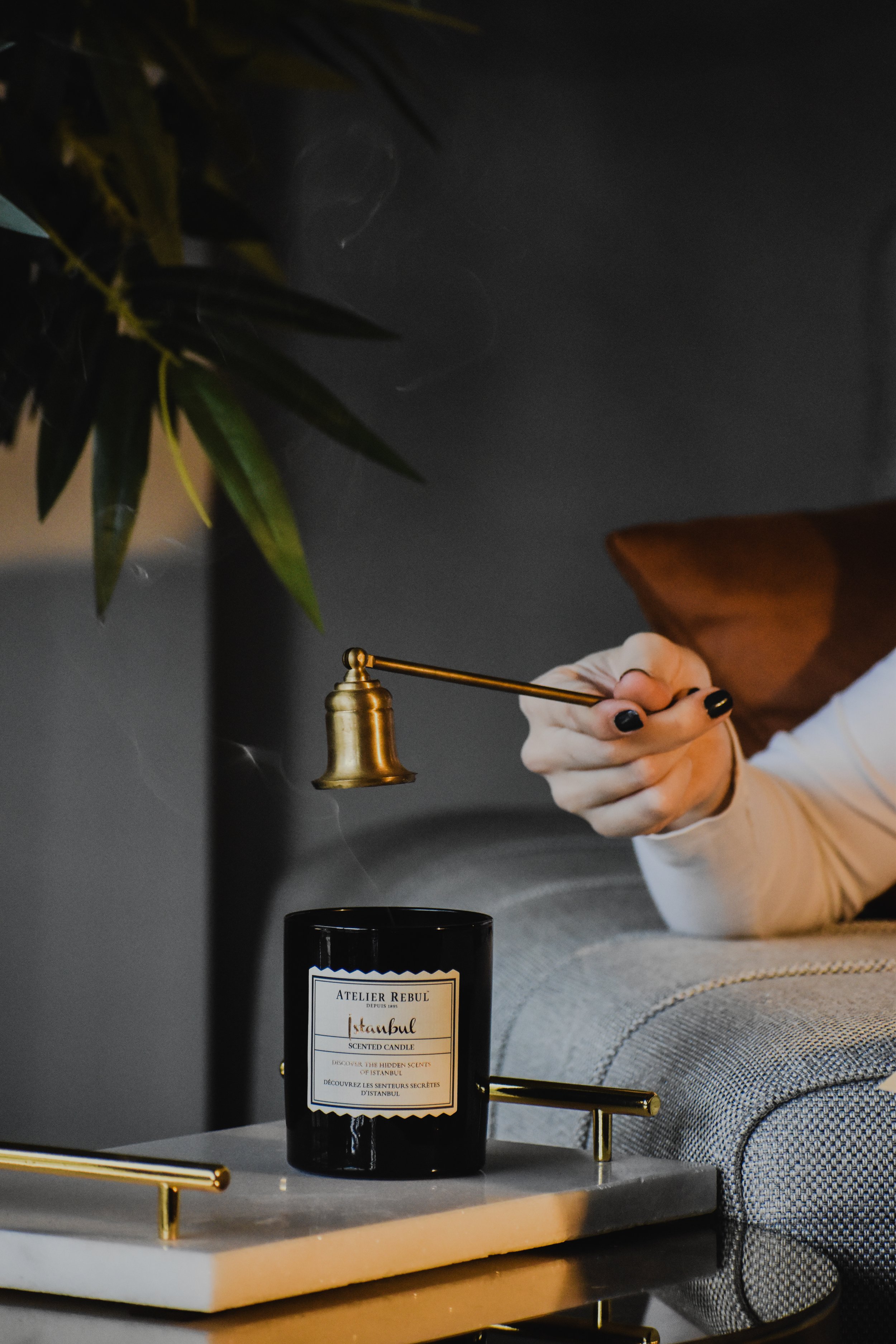 A person is holding a brass bell-shaped candle snuffer above a black candle with a label that reads 'Atelier Rebul Istanbul scented candle' placed on a white table with gold accents. The person is lying on a gray sofa with a brown pillow, with a green plant partly visible in the upper left corner.