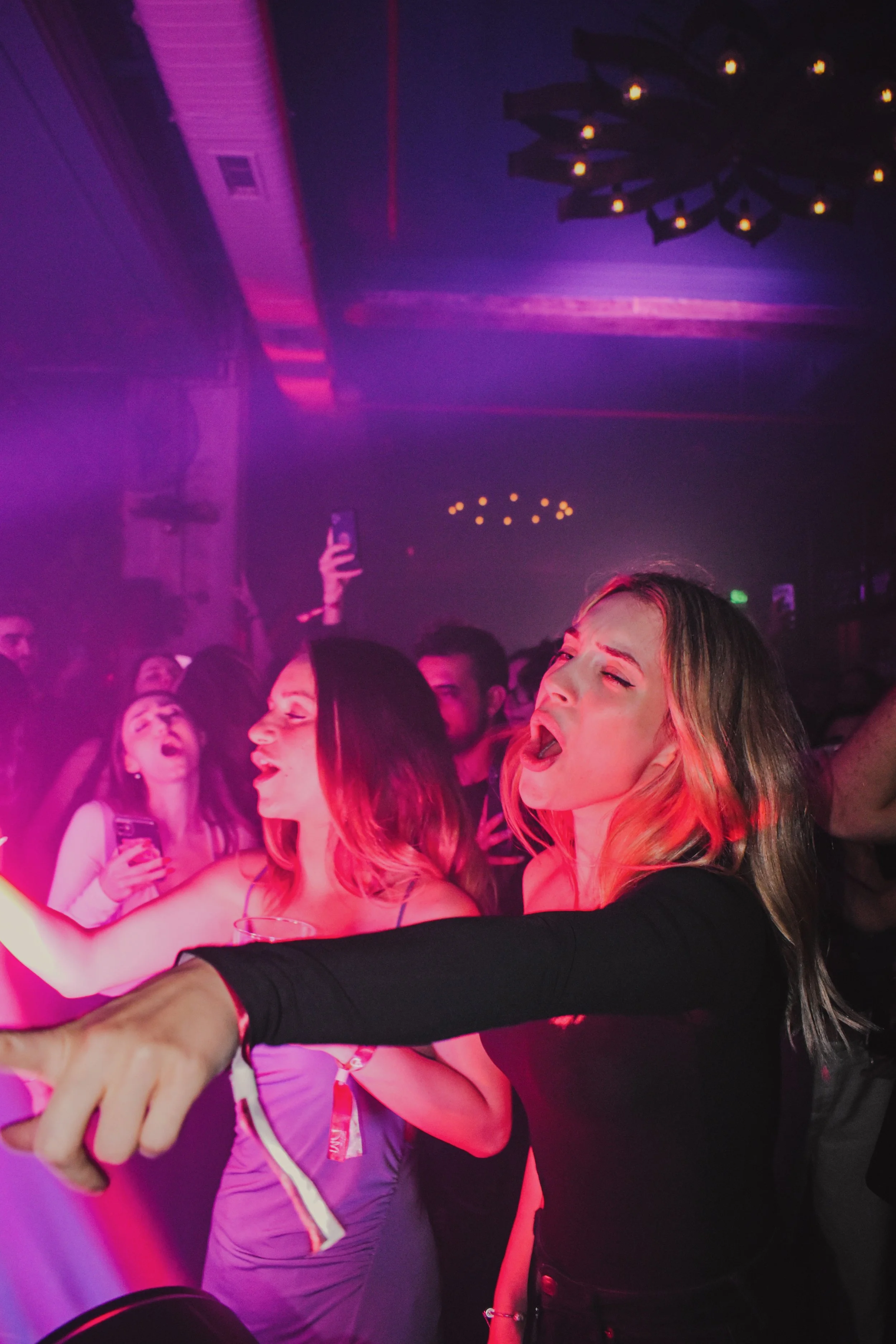 Group of young women and men dancing and enjoying themselves at a nightclub with colorful purple and pink lighting.