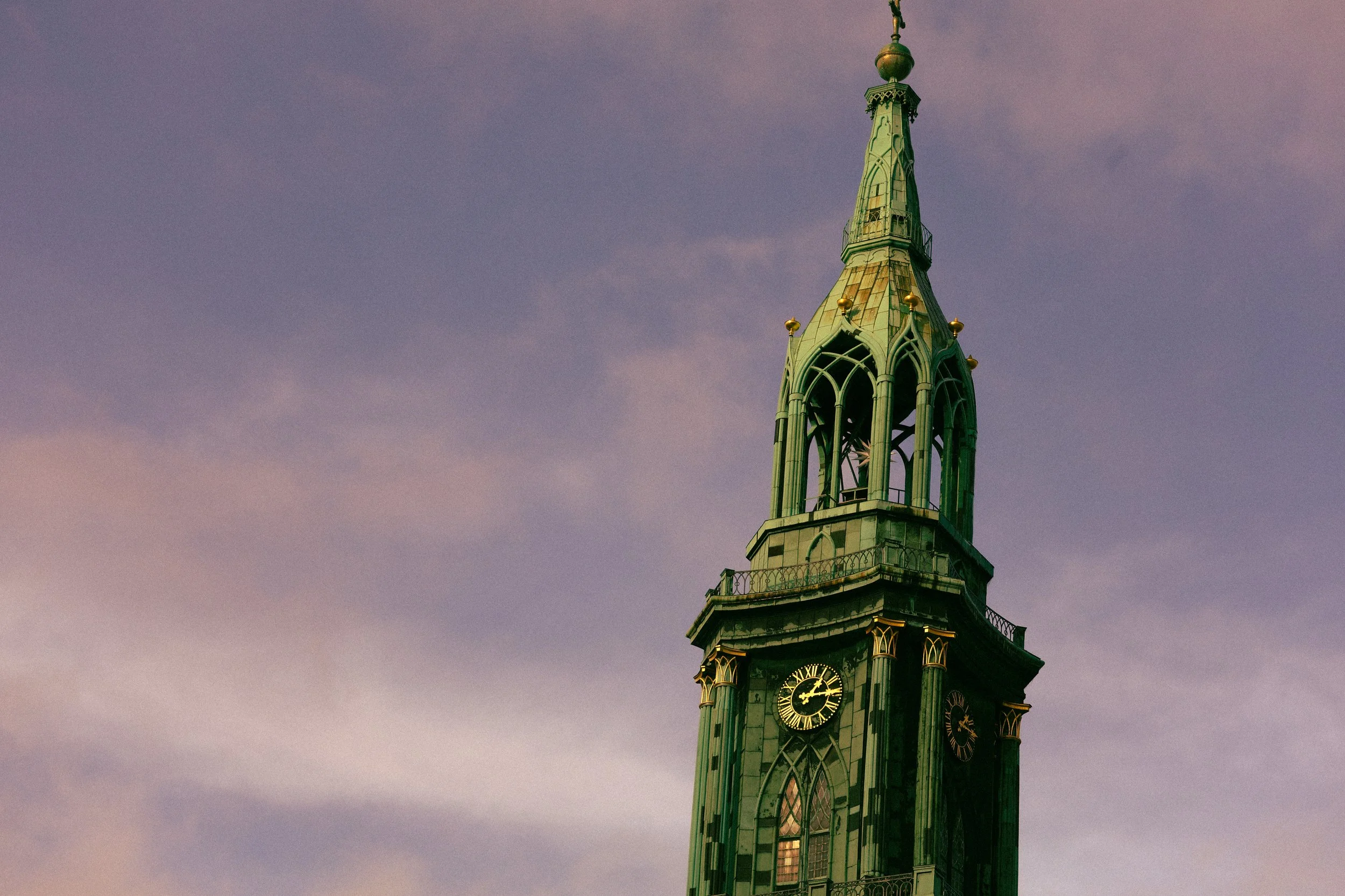 A green clock tower with a pointed spire against a pinkish-purple sky at sunset.