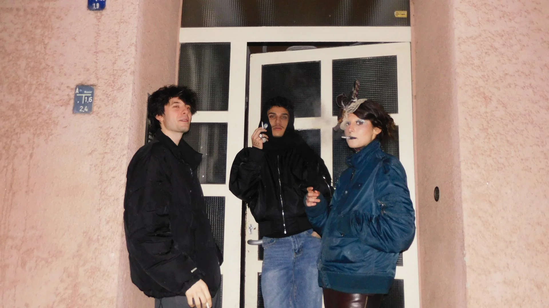 Three young adults standing outside near a door, dressed in vintage or retro clothing, with two of them smoking. The person on the left has dark, messy hair and is wearing a black jacket, the middle person has short dark hair and is wearing a black hoodie, and the person on the right is wearing a blue jacket and a decorative hat, smoking a cigarette.