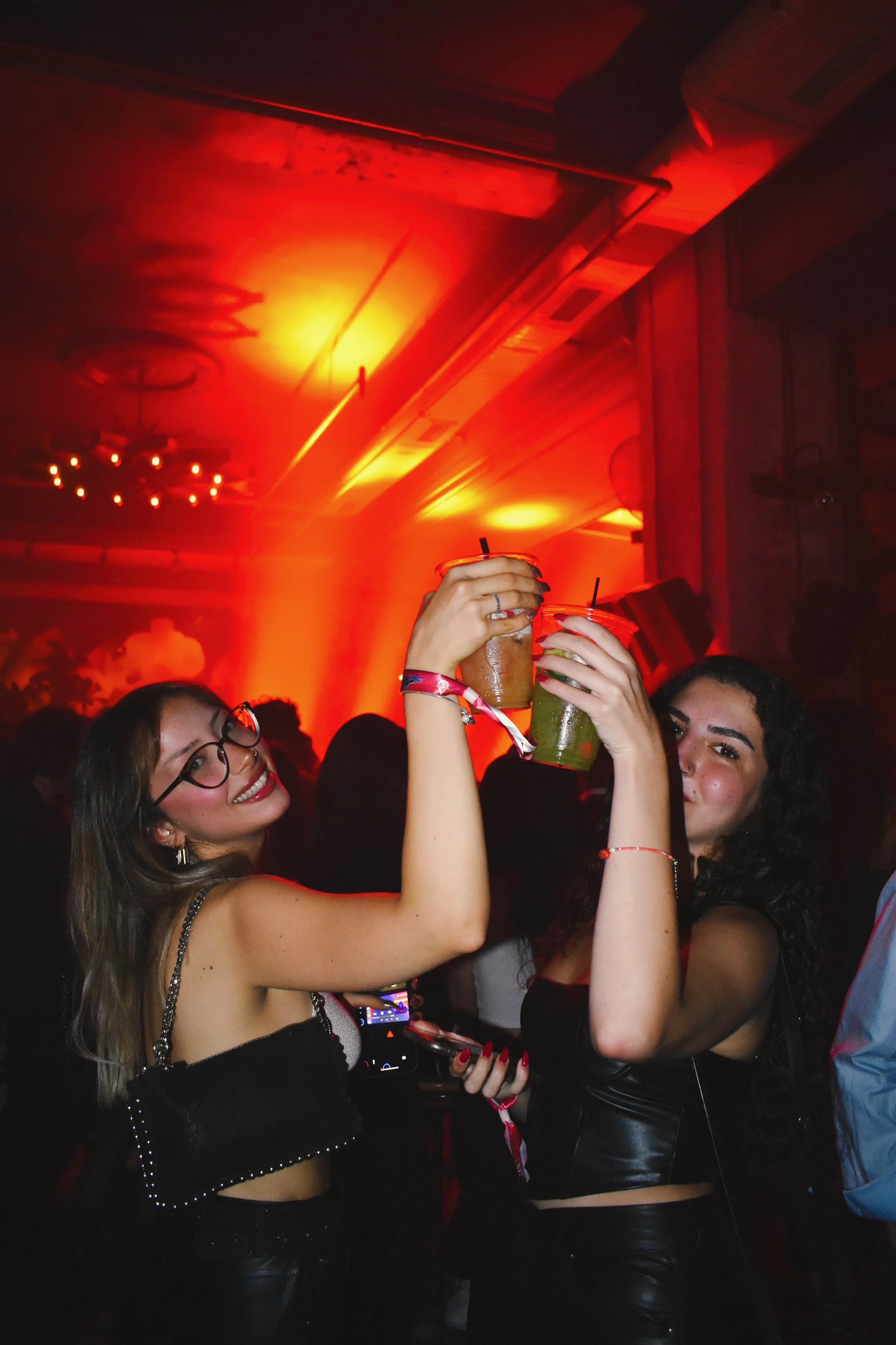Two women at a nightclub raising drinks and clinking glasses, with red and orange lighting, smiling and enjoying the party.