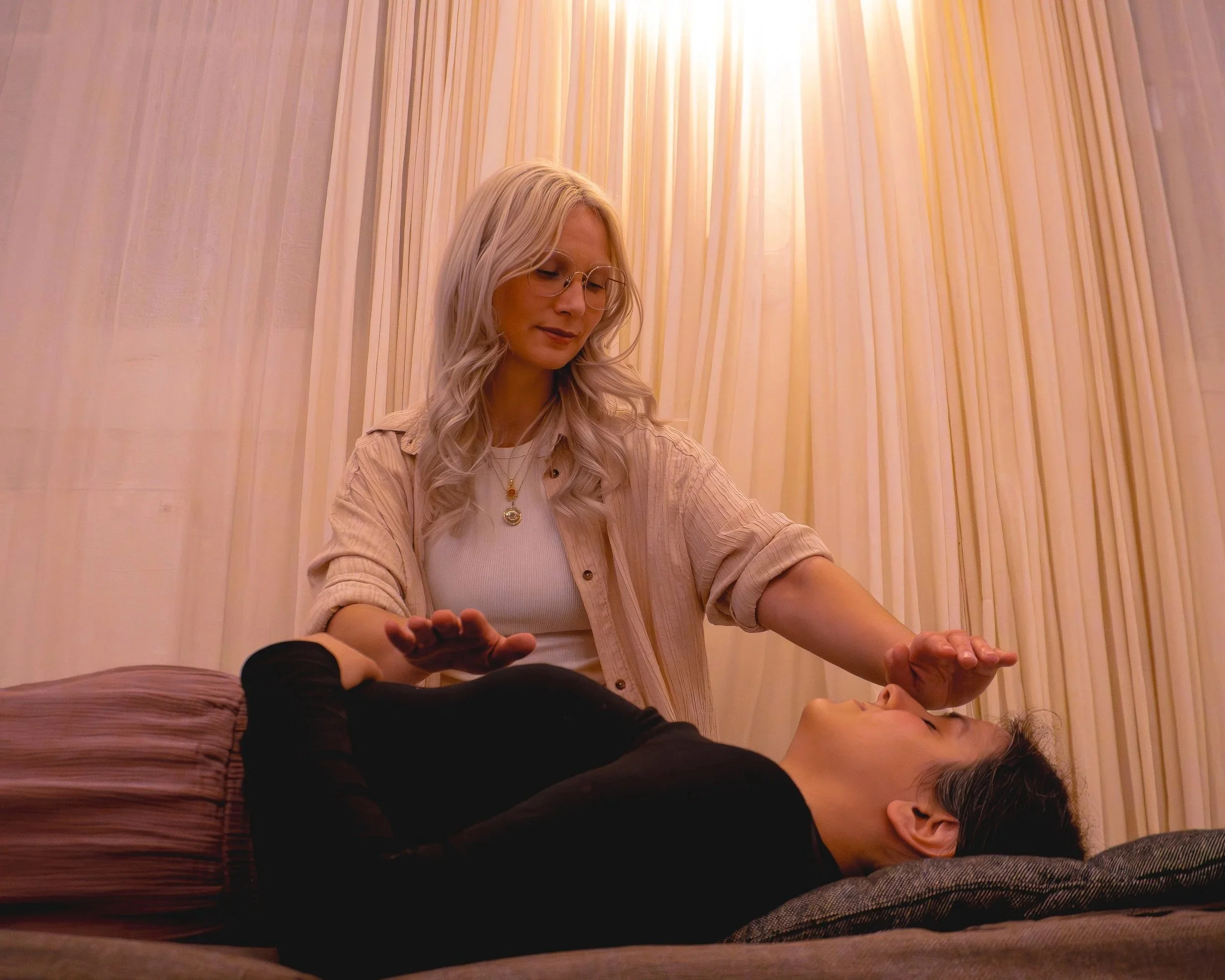 A woman with long blond hair wearing glasses and a beige shirt provides healing Reiki energy to a woman lying on a massage table, with her eyes closed and hand raised near her face, in a softly lit room with cream-colored curtains.