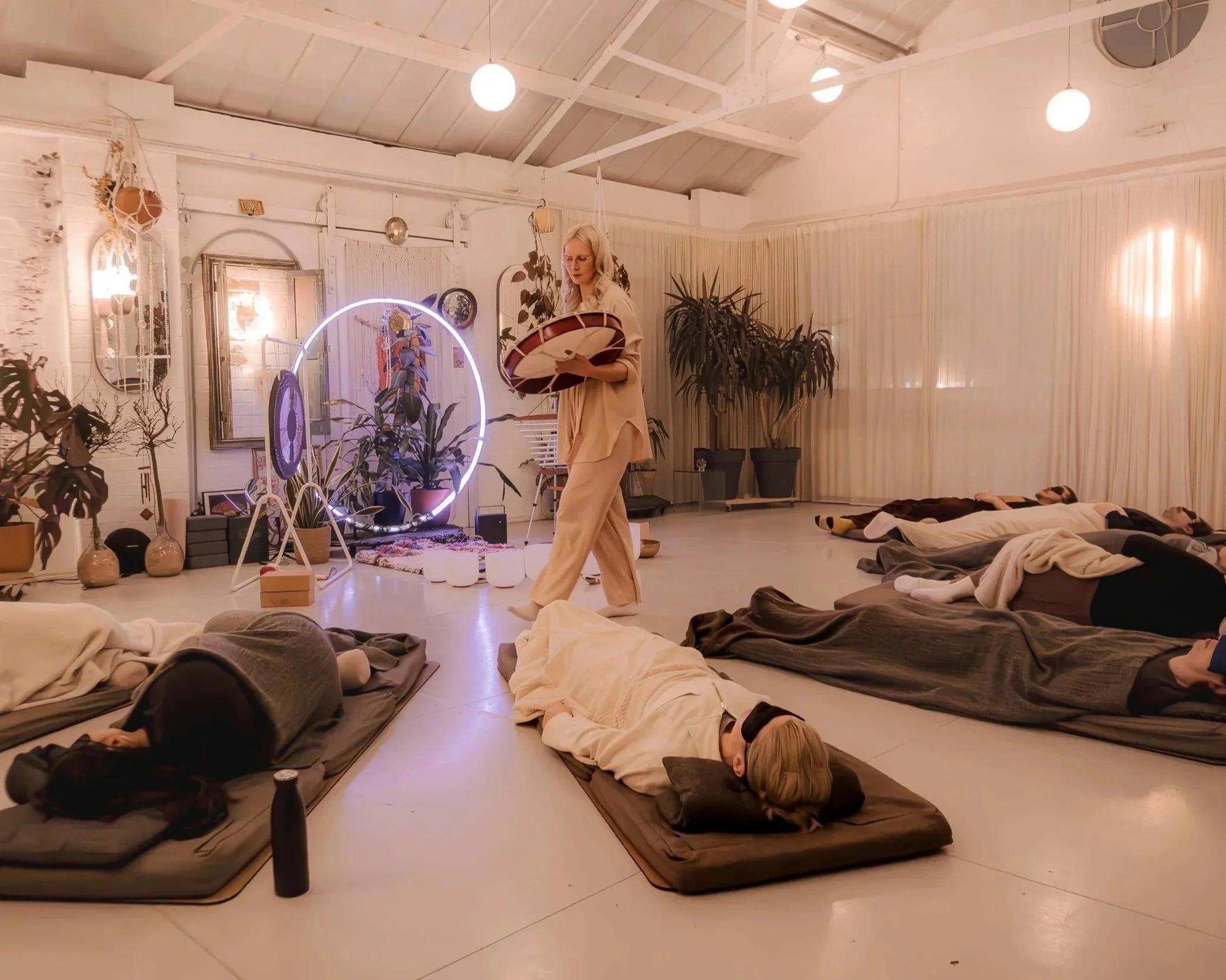 A group of people lying in reclined positions on mats, participating in a sound bath session in a warmly lit, decorated indoor space with plants and artistic decor, while a woman stands in the center playing a shamanic drum.