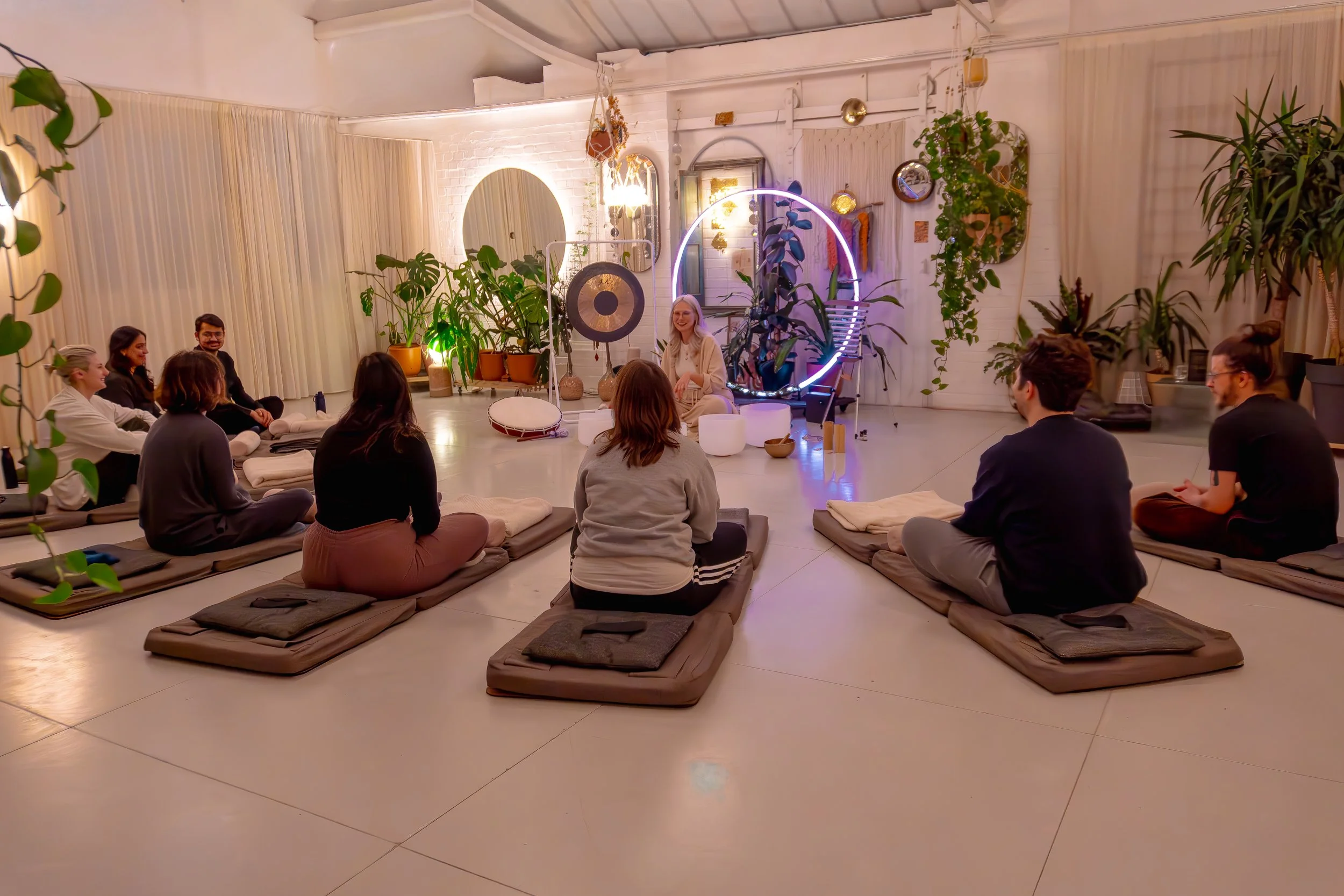 A group of people sitting on cushions in a circle, attending a sound bath in a cozy, plant-filled room with ambient lighting and decorative decor.