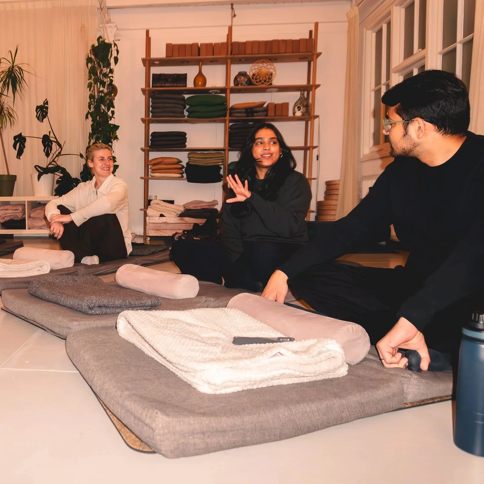 Three people sitting on yoga mats and cushions inside a room, engaging in conversation after a sound bath. Shelves with folded blankets and decorative items are in the background, and a water bottle is on the floor.
