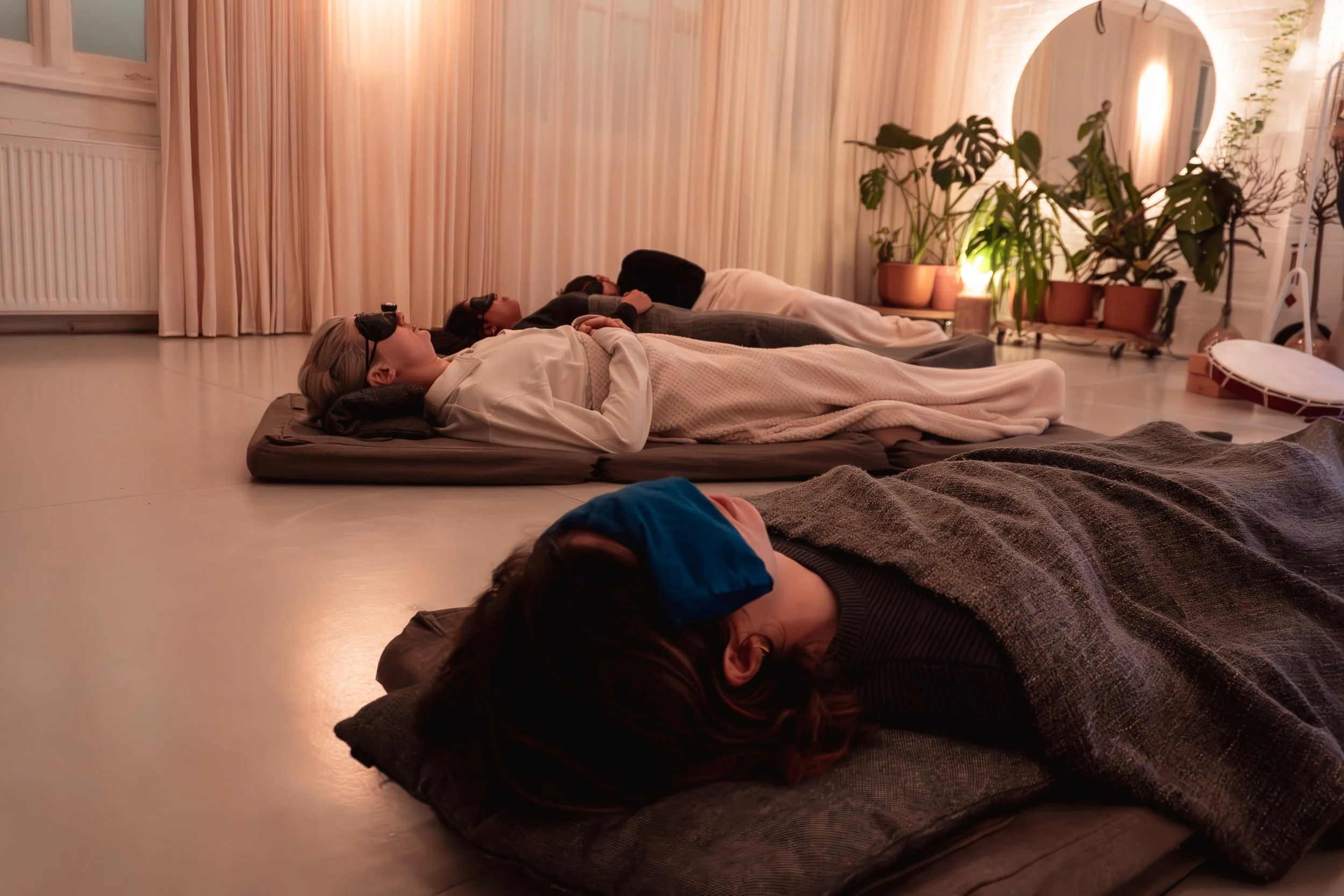 People lying down on pillows with eyes covered, participating in a sound bath session in a cozy, warmly lit room.