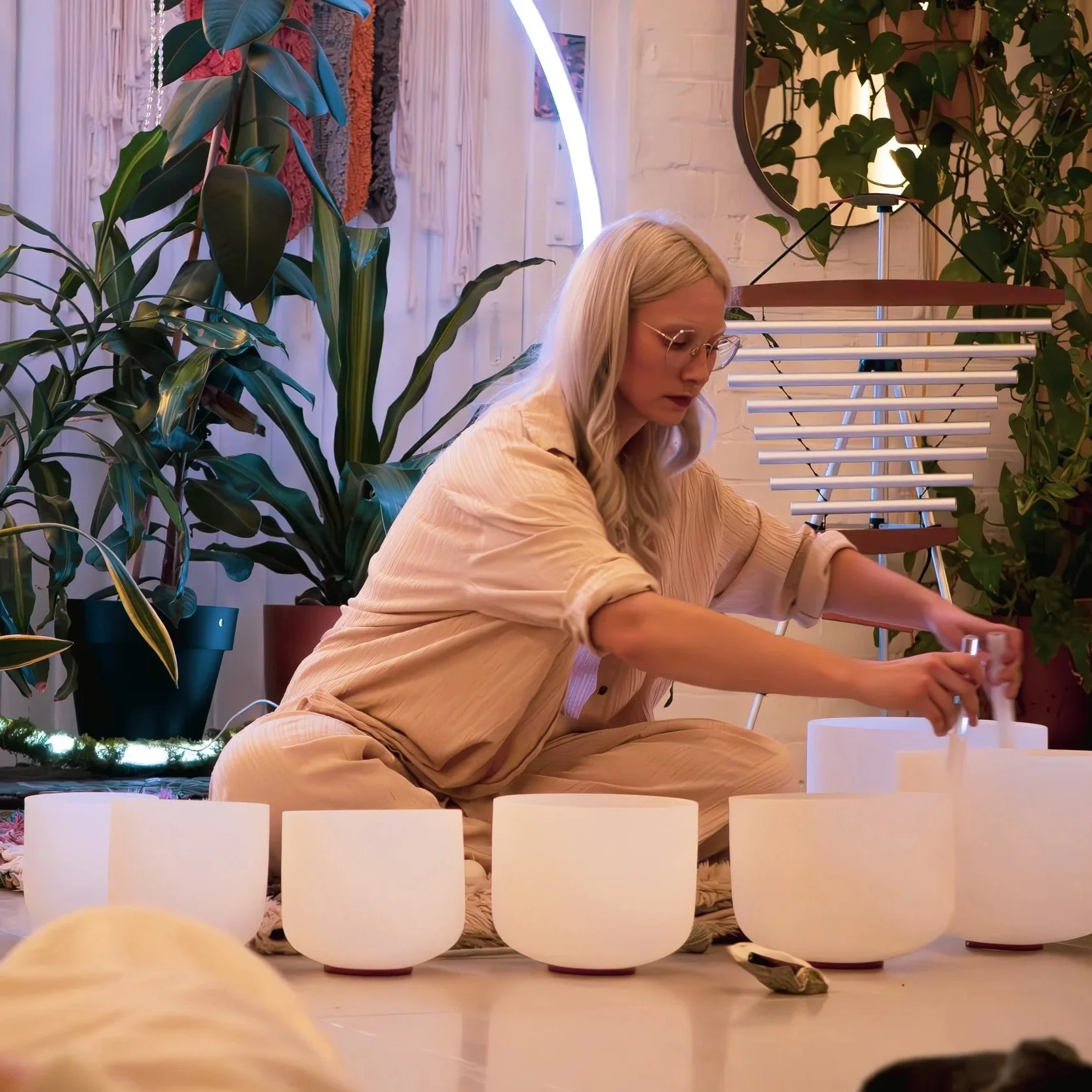 A woman with long blonde hair and glasses is sitting cross-legged on the floor, playing crystal singing bowls surrounded by green plants and decorative lights during a sound bath.