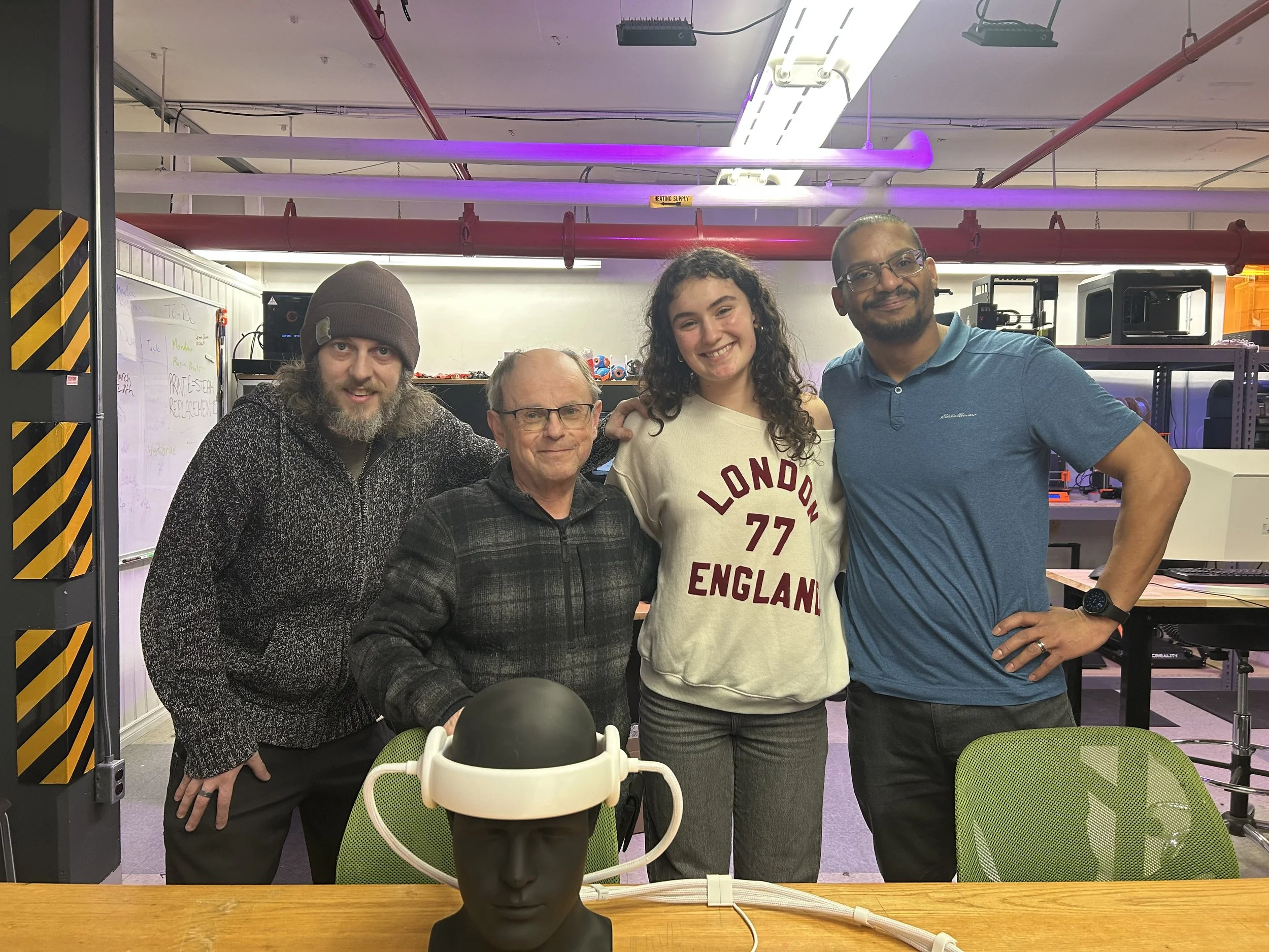 Four people standing in a room with technological equipment. A man with long hair and a beanie, a man with glasses, a woman with curly hair in a sweatshirt, and a man with glasses wearing a polo shirt. A mannequin head with a virtual reality headset is on the table in front of them.